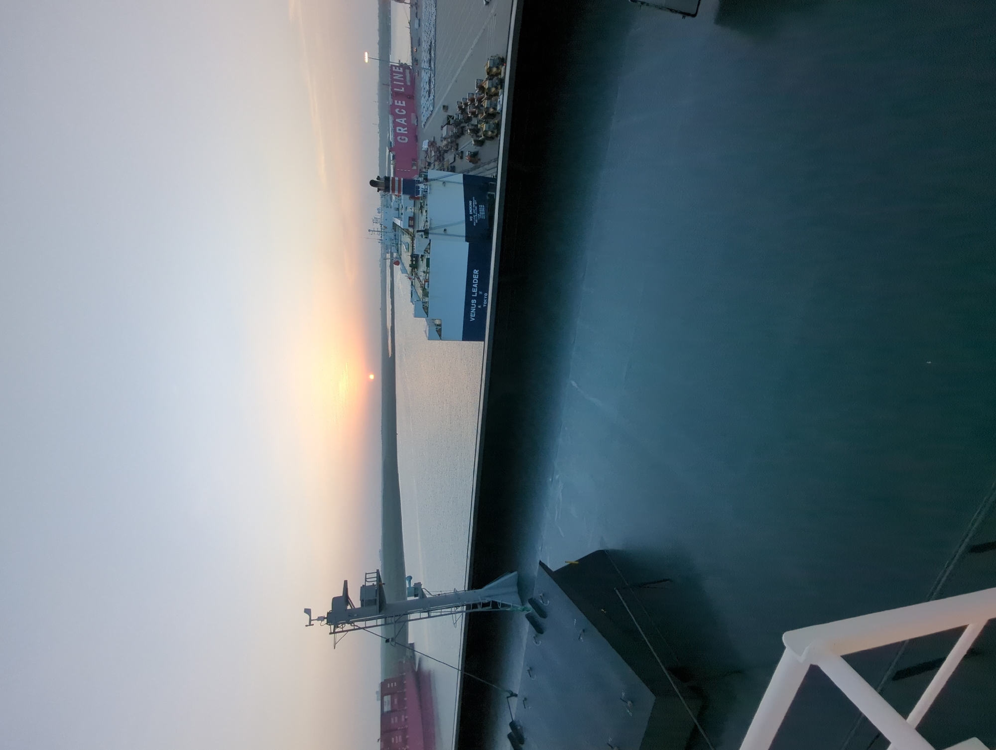 The image depicts a port scene at dusk in Hambantota, Sri Lanka. A large car carrier ship named "VENUS LEADER" is docked, laden with numerous vehicles, and another large pink ship labeled "GRACE LINE" is also visible. The sun is low on the horizon, casting a warm glow. The foreground shows the deck of a vessel, with a railing and a mast structure. The water is calm, reflecting the sky and the ships. The port appears to be a busy logistical hub.