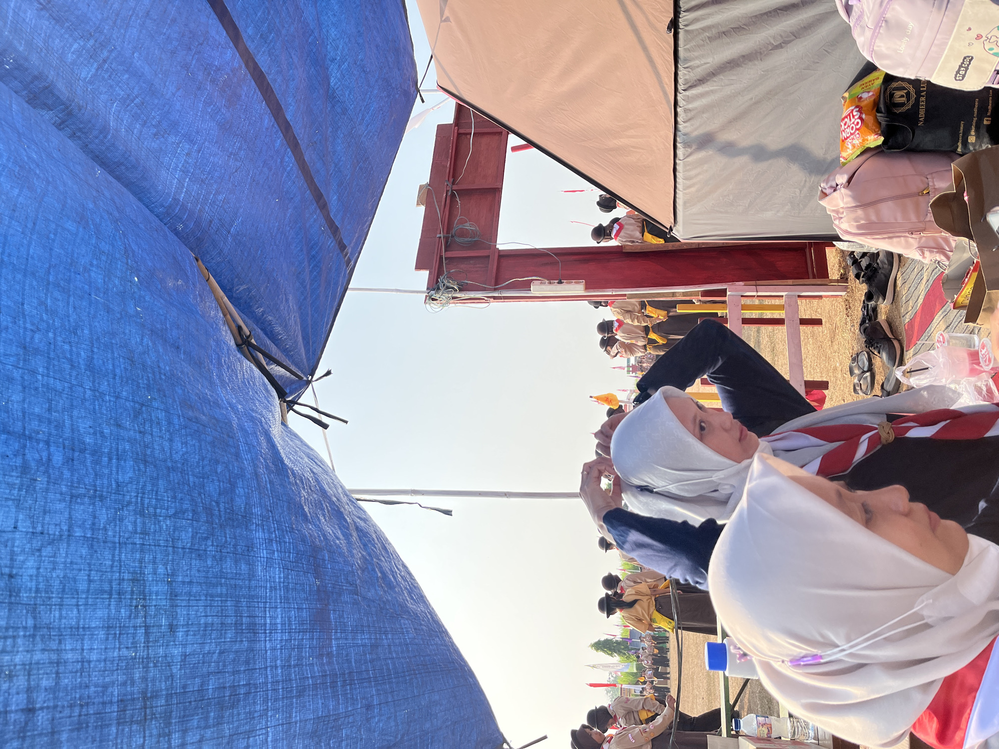 Two young women wearing white hijabs and scout scarves are in the foreground, preparing for an event. The woman on the right is adjusting her hijab, while the woman on the left looks down. Behind them, a group of scouts in uniform are lined up, possibly for an opening ceremony or parade. The scene appears to be outdoors, under a blue tarp, with a tent on the right and a red wooden archway in the background. The ground is covered with sand and scattered with shoes and bags. The event is likely a scouting jamboree or competition, possibly taking place in Gembong, Indonesia. The overall atmosphere suggests a sunny day and an organized outdoor gathering.