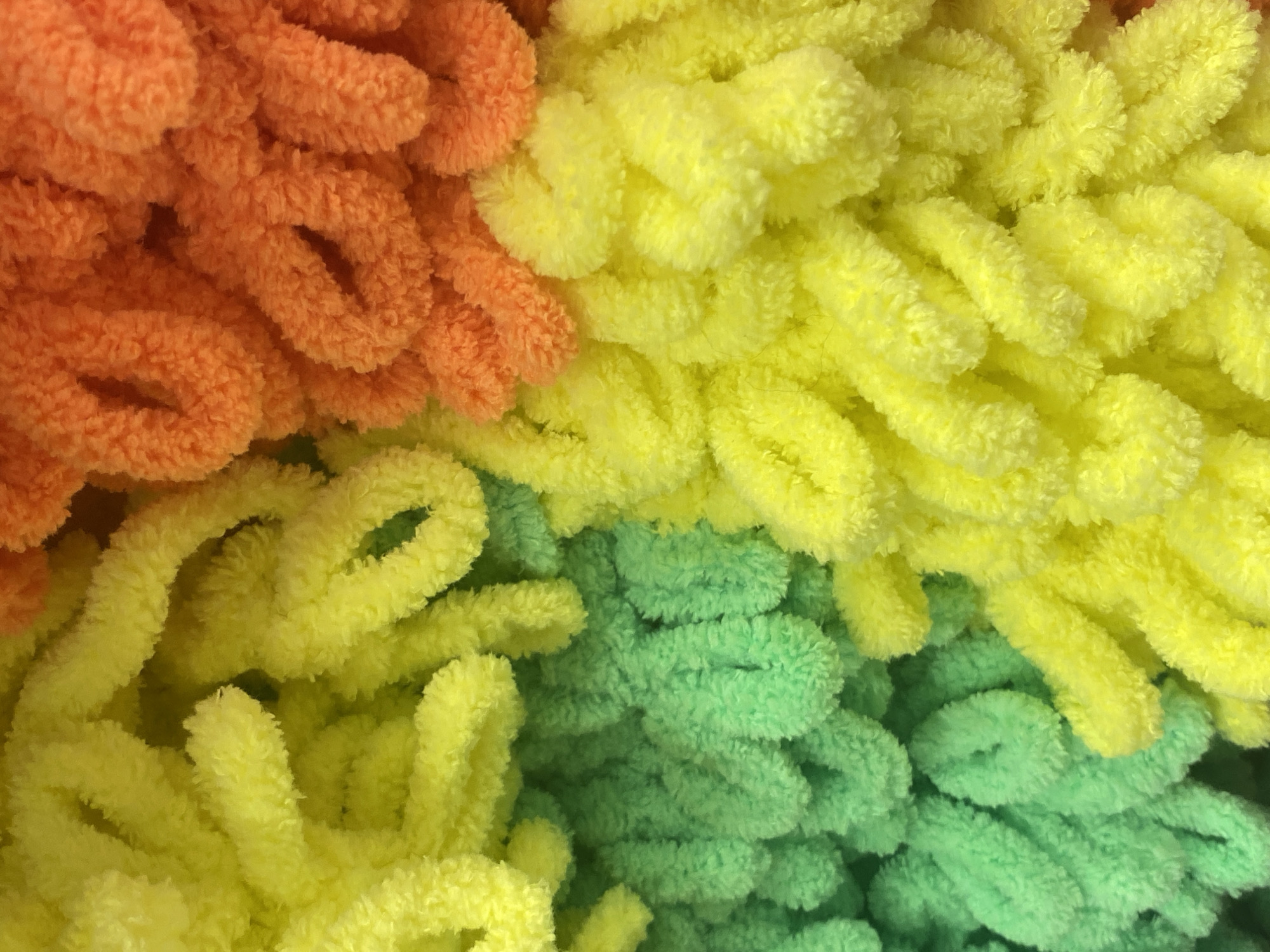 The image displays a close-up, abstract view of a fluffy, looped yarn in a gradient of colors. The yarn transitions from bright yellow on the left to a vibrant green in the middle, then to a warm orange on the right. The loops are densely packed, creating a textured surface. No specific location in Krasnodar, Russia, can be identified from this image. There are no people, objects, or actions depicted.