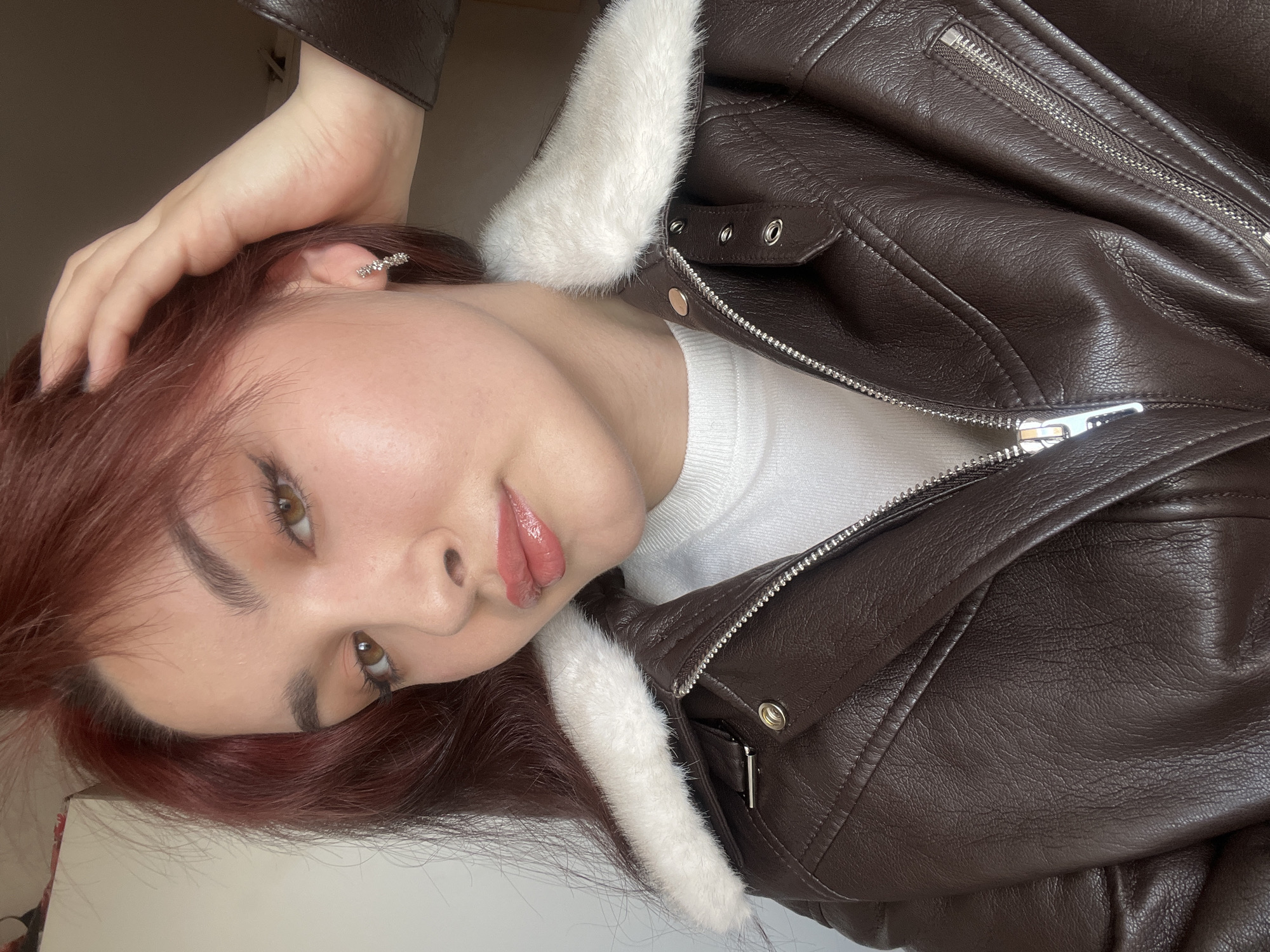 A portrait of a woman is presented in a sideways orientation. She has light skin, auburn hair, and light brown eyes. The woman is wearing a white sweater and a brown leather jacket with white fur trim. Her right hand is raised near her head, obscuring part of her hair. A silver earring is visible on her left ear. The background appears to be a plain, neutral color. There is no information in the image to suggest the location of Neftekamsk, Russia.