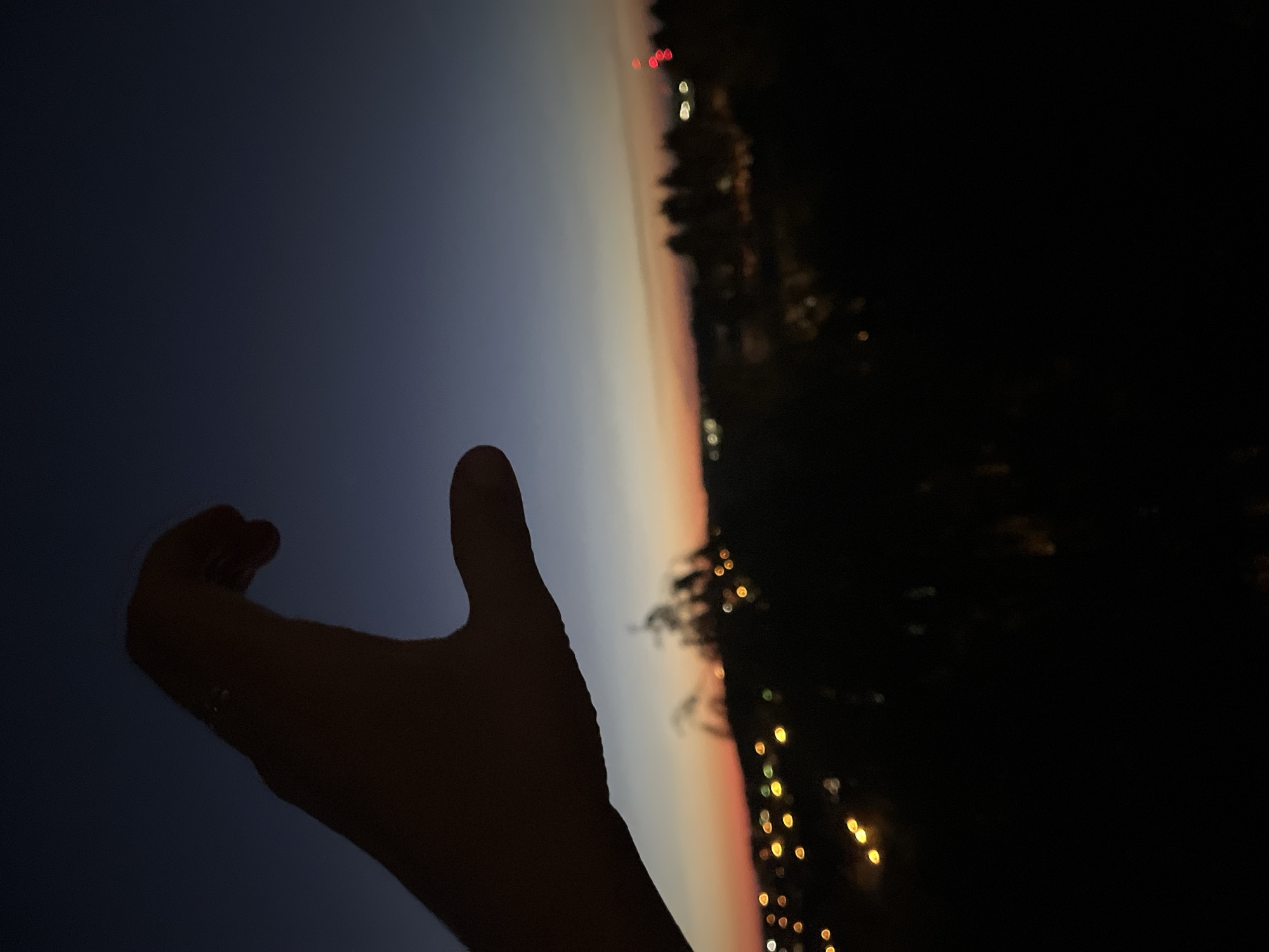 A silhouette of a hand is visible in the foreground, extended towards the right side of the frame. The hand is in a relaxed, open position, with the fingers gently curved. Beyond the hand, the scene depicts a twilight sky transitioning from a deep navy blue at the top left to a soft orange and pink hue closer to the horizon.
In the background, a dark silhouette of trees or foliage runs along the right edge of the image. Interspersed within the trees and in the distance are blurred, bokeh lights, suggesting a settlement or a gathering of lights. These lights are predominantly yellow and white, with a few red lights visible higher up.
The time of day is clearly dusk or twilight, given the gradient of colors in the sky and the presence of artificial lights. The overall mood is serene and contemplative, with the hand reaching out towards the fading light. There is no visible text in the image. The location is unknown, but the presence of trees and lights hints at a natural or semi-urban setting at the edge of a populated area.