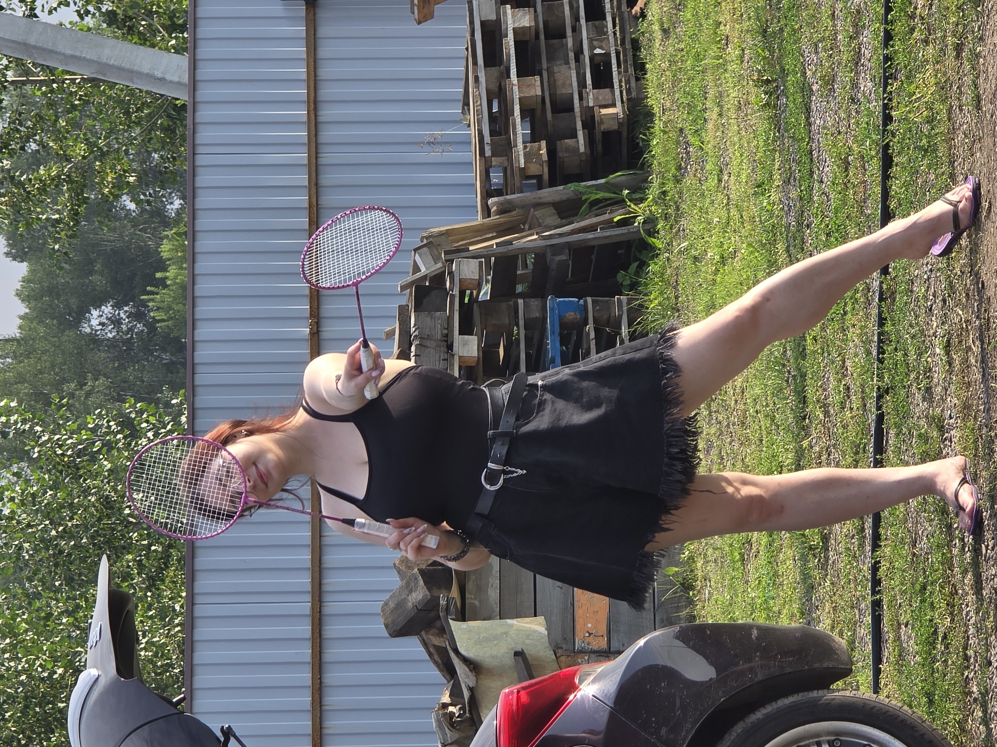 In an outdoor setting resembling a rural or suburban area near Ermolaevo, Russia, a young woman stands in front of a corrugated metal fence. She is wearing a black tank top and black denim shorts with a fringed hem, a black belt with a silver buckle and chain detail, and purple flip-flops. The woman is holding two badminton rackets, one in each hand, with the heads of the rackets positioned in front of her face. Her right leg is extended forward, and her left leg is bent at the knee. To her left, a portion of a dark-colored vehicle is visible. In the background, there are stacks of wooden pallets and various green foliage, including trees. The ground is covered with grass and gravel.