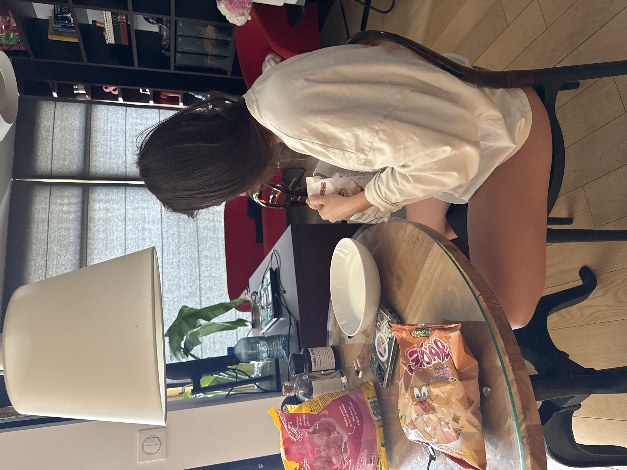 A person with long brown hair is seated at a round table, facing away from the camera and slightly bent forward. They are wearing a white long-sleeved shirt and appear to be eating from a packet of "FOAR" cereal pillows. The table is cluttered with various items, including a white bowl, medicine bottles, a bag of "SHAKE" cereal pillows, a plastic bottle of water, and some small electronic devices. A lamp with a white shade is situated on the left side of the frame. Behind the person, a black chair is visible, along with a red chair and a black shelf filled with books and various items. A window with blinds is behind the table and lamp. The location appears to be an indoor residential setting, possibly in an apartment. There is no indication in the image that the location is in Warsaw, Poland.