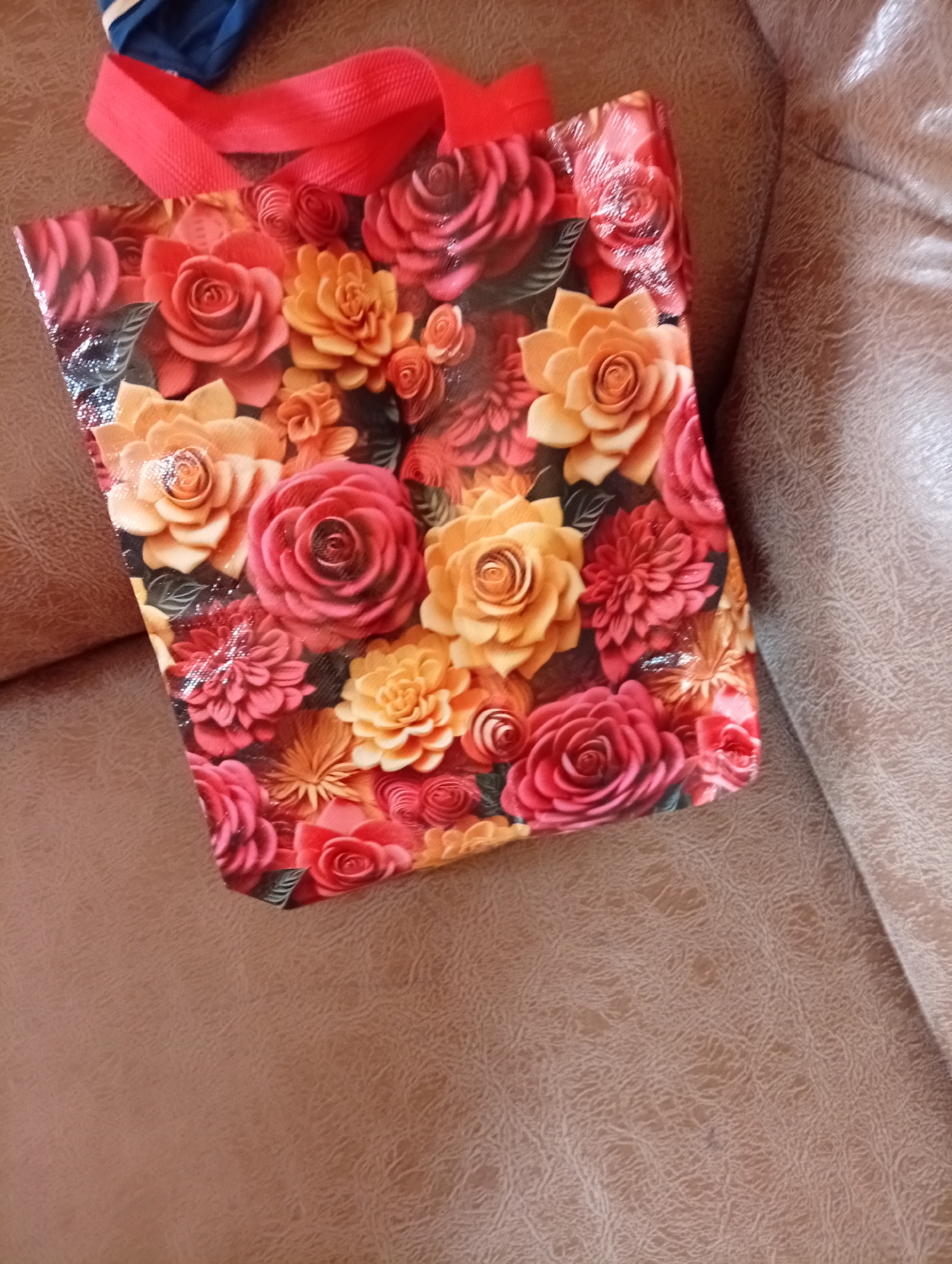 The image shows a close-up, high-angle shot of a floral gift bag with red straps resting on a brown textured surface, likely a sofa or upholstery. The bag features a vibrant pattern of roses and other flowers in shades of red, pink, and orange, with some areas appearing to have a glossy or metallic finish. A small portion of a blue object is visible in the upper left corner, partially obscured by the red strap. The lighting suggests an indoor setting. The image does not depict any specific location in Gura Top, Nigeria, or any people.
