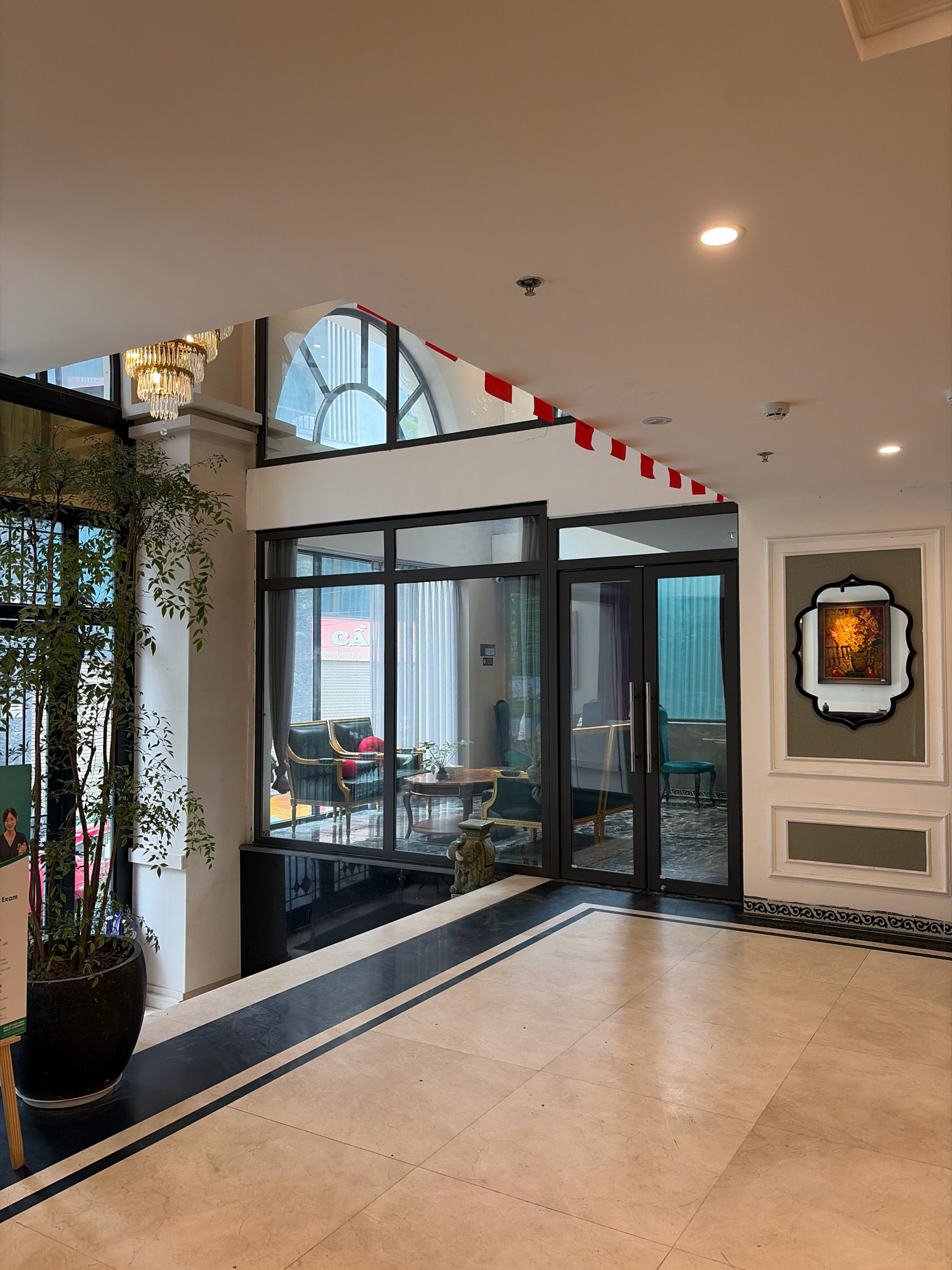 The image shows an interior view of what appears to be a hotel lobby or a high-end commercial space in Hanoi, Vietnam. The floor is tiled with large, light-colored marble tiles, bordered by a black and white striped pattern. To the left, a tall potted plant with lush green leaves stands next to a glass door with a metal grate. A woman is partially visible in the background on the left, standing behind a green sign.
Further into the space, a large glass-enclosed area reveals an interior seating arrangement with dark green velvet armchairs and a round wooden table. Beyond this, through another set of glass doors, a glimpse of what looks like a lounge or another room can be seen. Above, a decorative chandelier hangs from the ceiling, and red and white triangular flags are strung across the upper part of the frame. A framed artwork is displayed on the wall to the right, featuring a still life of flowers. Recessed ceiling lights and a smoke detector are also visible. The architecture suggests a modern yet elegant design, possibly incorporating influences from Vietnamese aesthetics given the location.