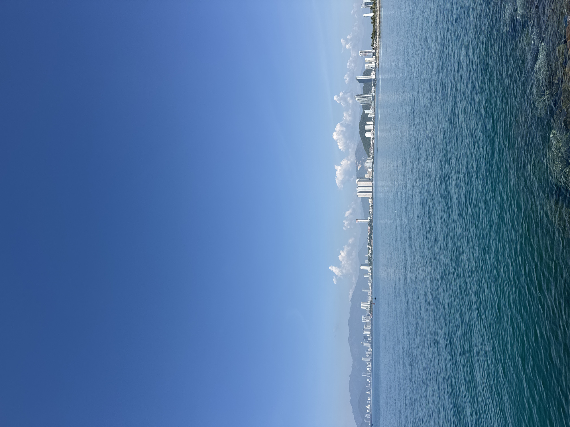 The image shows a wide, calm expanse of blue-green ocean in the foreground, with gentle ripples on its surface. In the middle ground, a city skyline stretches across the horizon, characterized by numerous tall, modern buildings of varying heights and architectural styles, predominantly white. Behind the city, a range of low, green mountains forms a backdrop, with some haze visible. The sky above is a clear, bright blue, with a few scattered white clouds. The lower right corner of the image shows the edge of a rocky shoreline. The scene is a panoramic view of a coastal city. Based on the urban development and the prominent mountain range, the location is likely the city of Nha Trang, Vietnam, specifically the coastal area of Bac Nha Trang Ward.