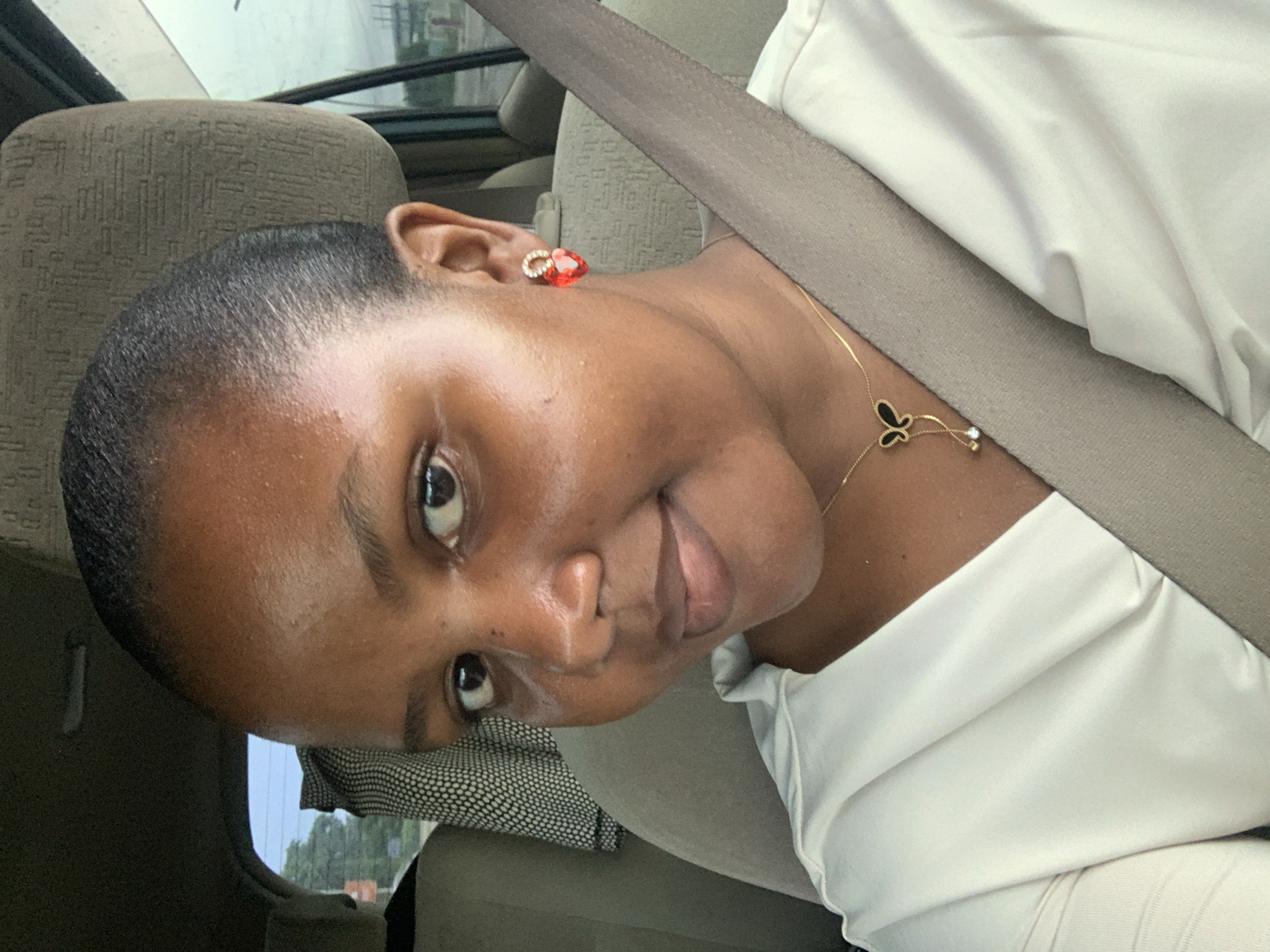 A woman is in the passenger seat of a car. She is wearing a white shirt and a seatbelt. She has a gold necklace with a butterfly pendant and red earrings. The car's interior is gray. The background is blurry and indistinct. The location cannot be determined from the image.