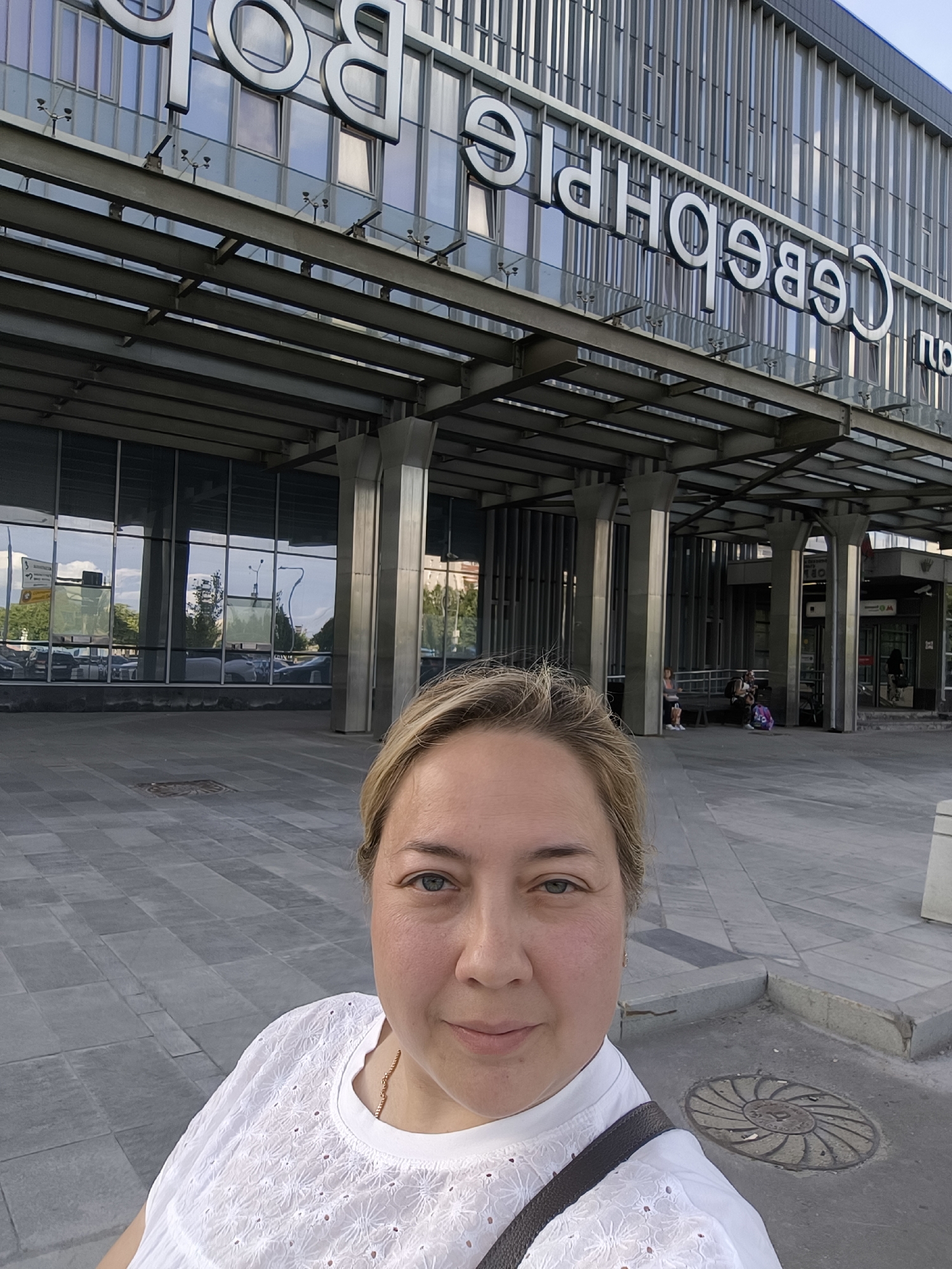 A woman with blonde hair and blue eyes is taking a selfie outdoors in front of a building with large glass windows and metal signage. She is wearing a white, embroidered blouse and a dark strap across her chest. The building has a modern architectural design with vertical metal slats and large, reflective windows. In the background, people can be seen sitting on benches and walking. The ground is paved with large gray tiles. The signage on the building appears to be in Cyrillic.