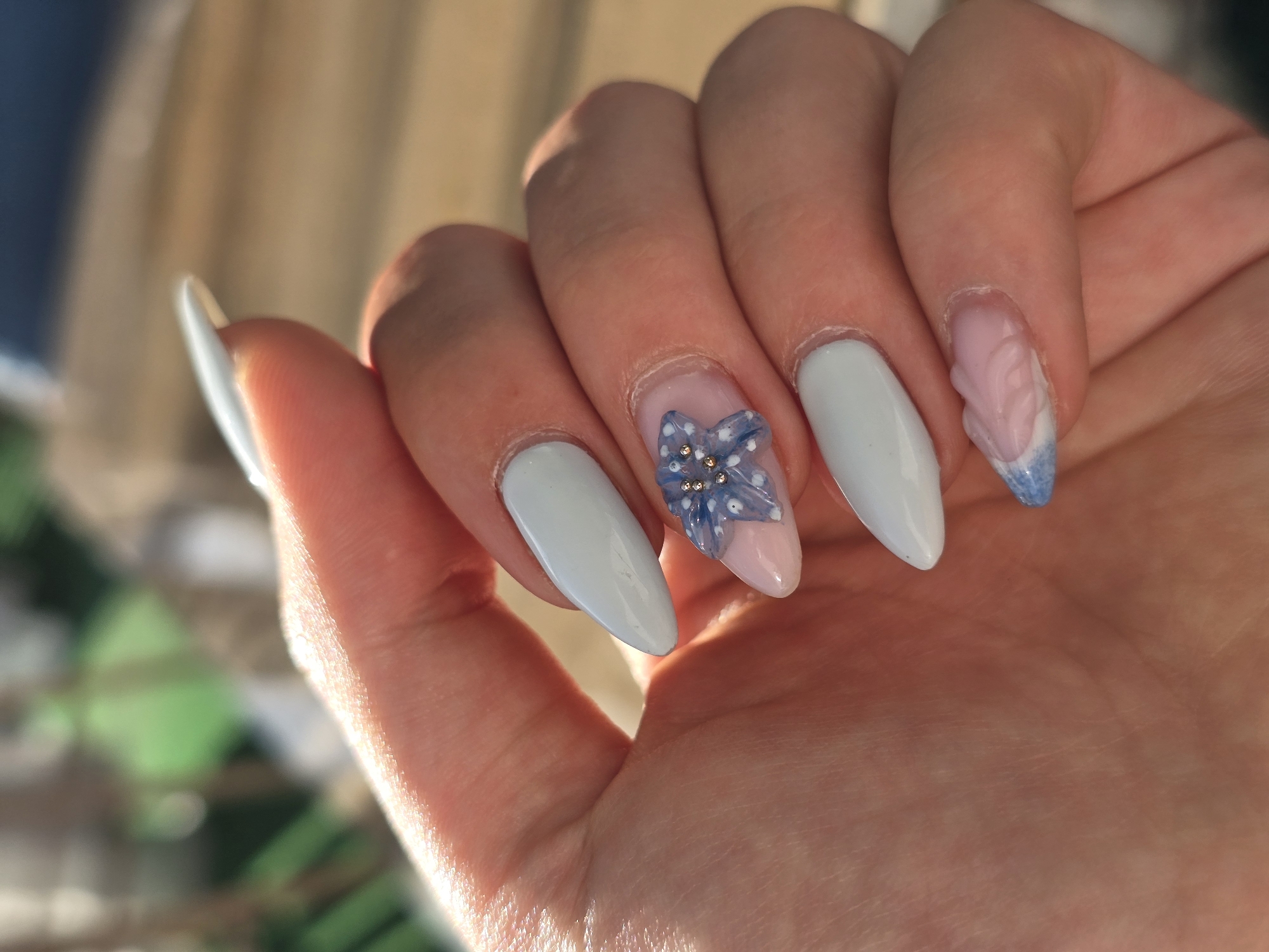 The image displays a close-up of a person's left hand, meticulously manicured and brightly lit by natural sunlight, suggesting daytime. The hand is gently curved, presenting five long, pointed nails.
Four of the nails (thumb, index, middle, and ring fingers) are painted in a solid, opaque light blue or white shade. The ring finger nail features a prominent three-dimensional blue flower design, adorned with small white dots and centered with several tiny golden rhinestones. This nail also has a translucent pink or nude base beneath the flower. The little finger nail also utilizes a translucent pink/nude base, but instead of a flower, it showcases a raised, textured, wavy or feather-like pattern in a matching pink hue, which transitions into a soft blue gradient at the tip.
The background is softly blurred, showing warm, light brown and beige tones, possibly from a wall or wooden surface, with hints of out-of-focus green elements, suggesting either an outdoor setting or a bright indoor space near foliage. The lighting casts warm highlights on the skin and nail surfaces. There is no visible text. The image focuses entirely on the detailed nail art, implying the activity of showing off a freshly completed manicure. While the location context is Burgas, Bulgaria, no specific visual cues in the image corroborate this.