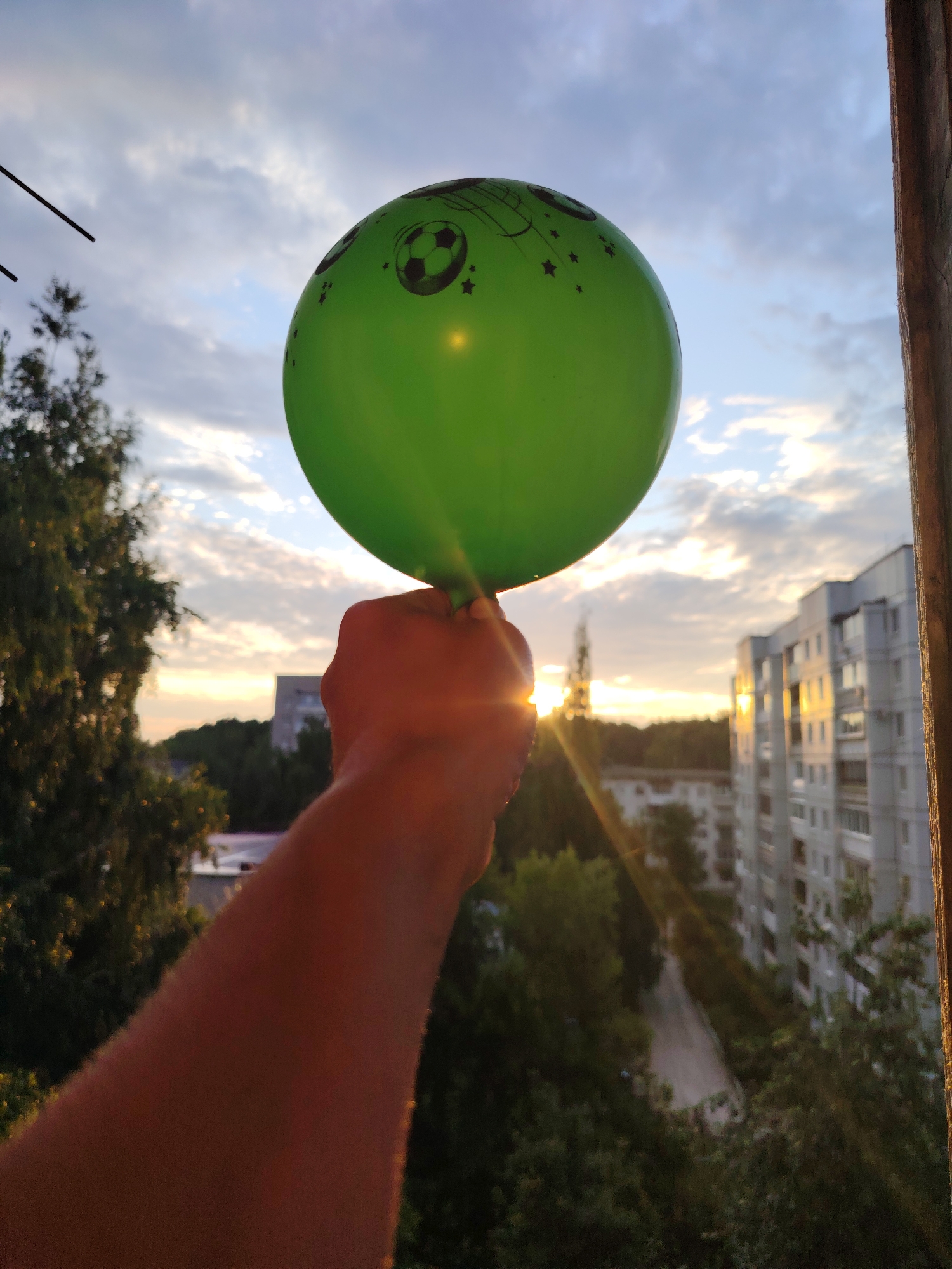 The image depicts a close-up view of a person's arm and hand holding a green balloon against a sunset sky. The balloon is decorated with small black stars and a soccer ball design. In the background, there are apartment buildings, trees, and a sliver of the Kama River visible, indicating the location is the city of Chaikovsky, Russia. The sun's rays are piercing through the clouds and the balloon, creating a lens flare effect. The scene appears to be captured from a balcony or window overlooking a residential area.