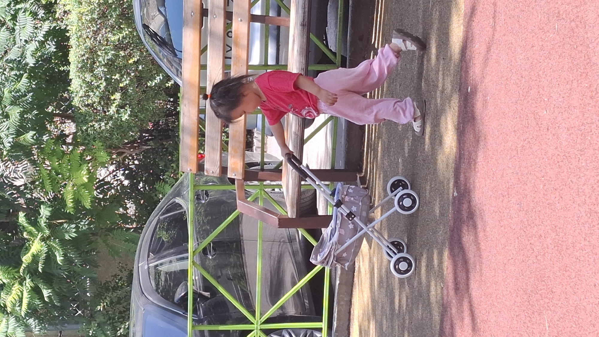 A young child of East Asian descent, likely a toddler, is seen pushing a toy stroller on a playground with a pink, textured surface. The child is wearing a bright pink t-shirt and pink pants, and is also wearing light-colored sandals. The child has dark hair pulled up in a small ponytail. The toy stroller is white with a grey and white patterned fabric basket adorned with star shapes. The stroller has four wheels. In the background, there is a wooden bench. Behind the bench, there are parked vehicles, including a dark-colored sedan. Lush green trees and foliage are visible, suggesting an outdoor urban setting. The lighting indicates it is daytime, with shadows present on the playground surface, indicating the sun is shining. The location is identified as the city of Shymkent, Kazakhstan.