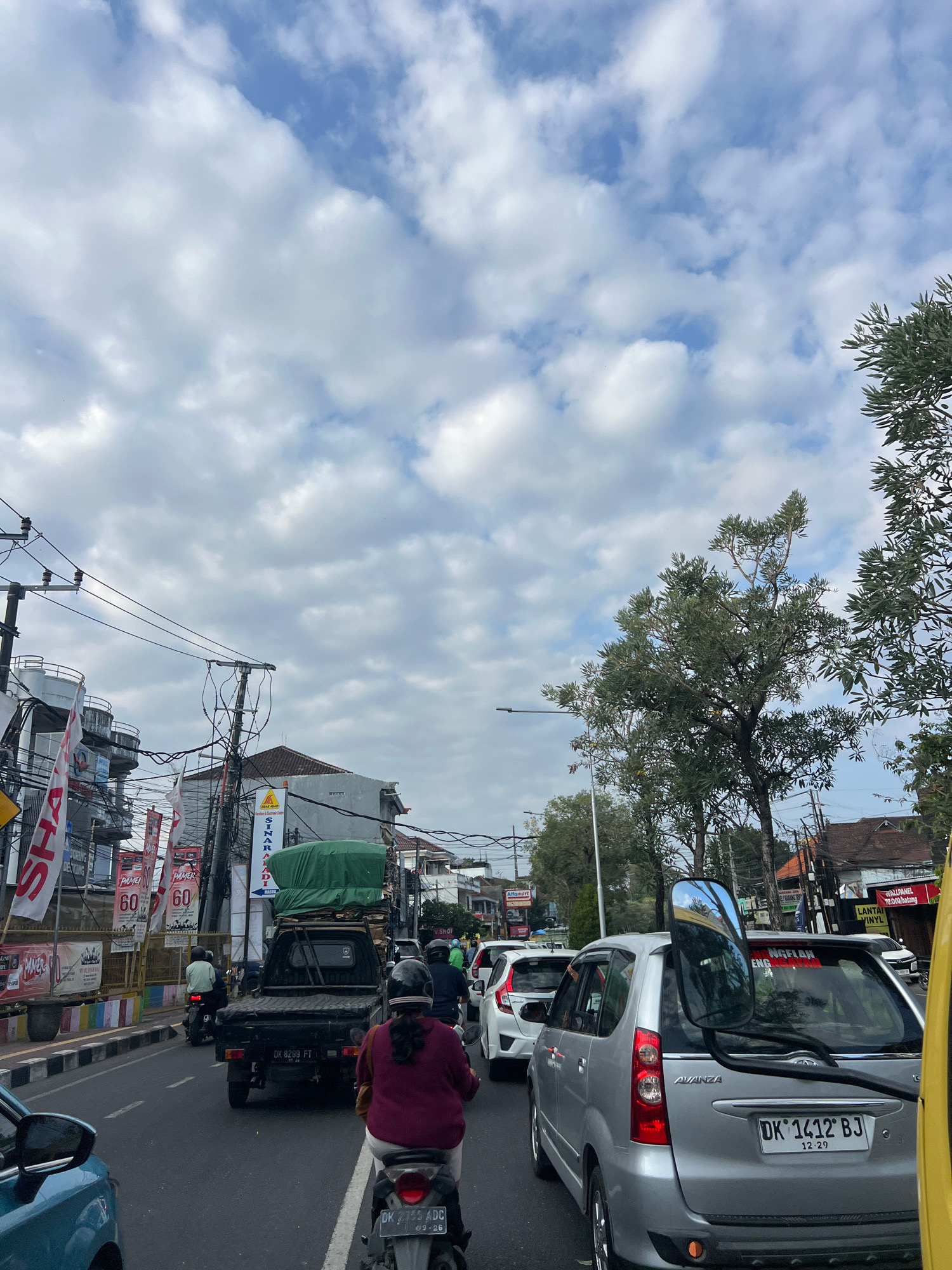 This is a medium-shot, eye-level photograph taken on a cloudy day in Denpasar, Indonesia, capturing a busy street scene with traffic congestion. The foreground is dominated by the backs of motorcycles and cars, indicating a traffic jam. On the right, a silver minivan with a large side mirror is prominently visible, its license plate reading "DK 1412 BJ". To its left, other vehicles are stalled. In the center of the frame, a black pickup truck loaded with green-covered goods is stopped, with motorcycles and cars behind it. On the left side of the road, buildings with various signs and utility poles with numerous wires are visible. The sky is filled with scattered white clouds against a pale blue backdrop. The overall impression is one of a typical urban environment experiencing moderate traffic flow under overcast conditions.