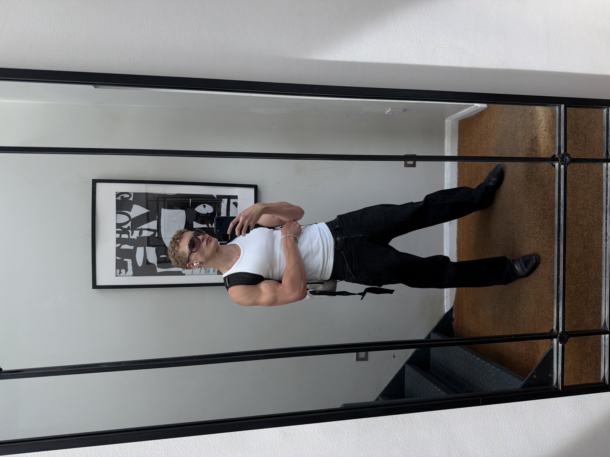A man with curly blonde hair, wearing sunglasses, a white tank top, black jeans, and black shoes, stands in front of a full-length mirror. He is holding a phone in his right hand, taking a selfie. The man is wearing white earbuds and a silver chain necklace. He also has a black backpack on. The mirror reflects a staircase with metal steps to his left, a framed black and white abstract artwork on the wall behind him, and a brown rug on the floor. The scene appears to be indoors, possibly in a home or a studio, with white walls and a grey accent wall. There is no discernible indication that the location is in London, United Kingdom.