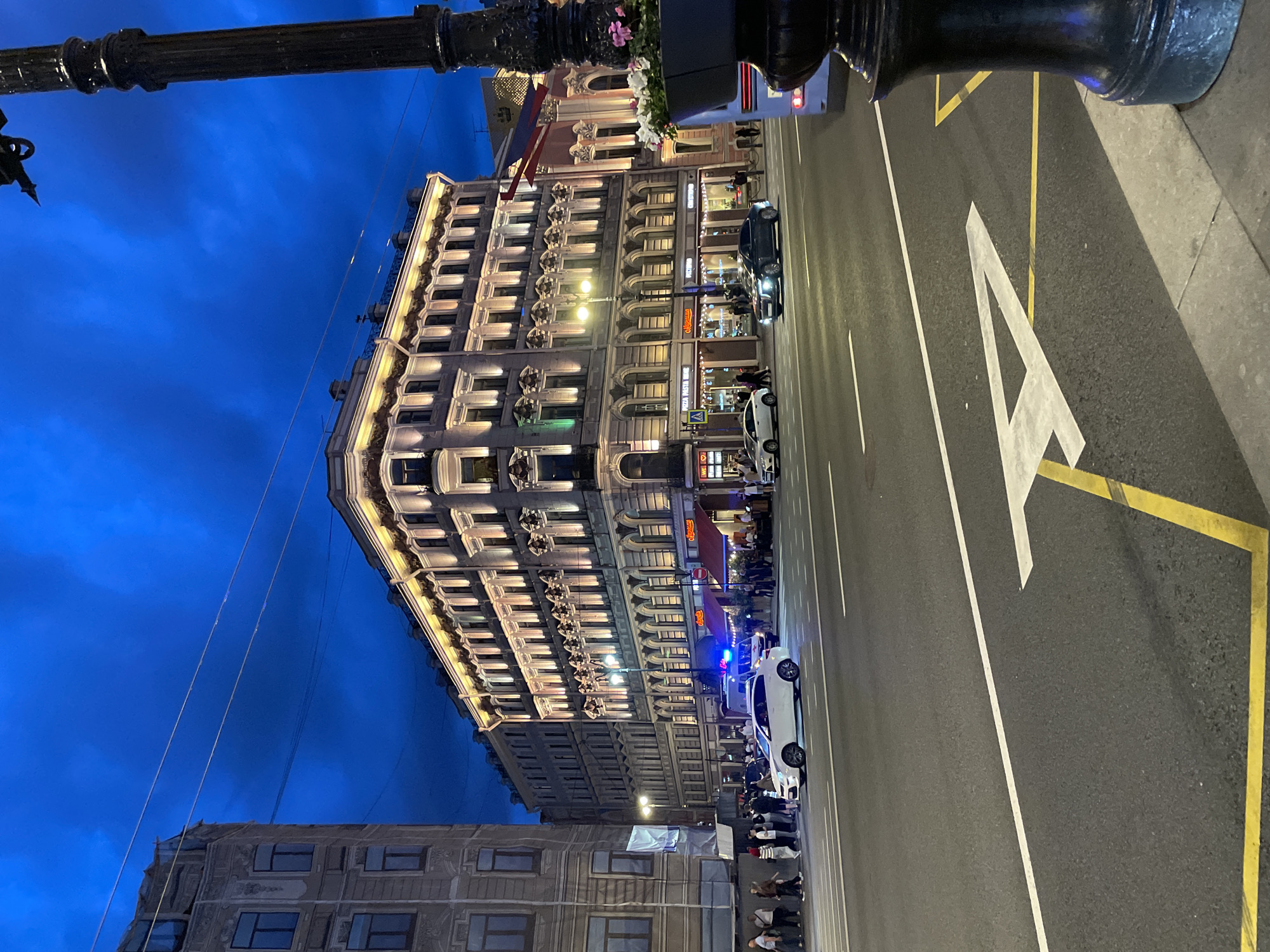 This image captures a nighttime urban scene on Nevsky Prospekt in Saint Petersburg, Russia.
The foreground features a wide, asphalt road marked with white solid and dashed lines, indicating traffic lanes. A large white arrow points forward in the rightmost lane, complemented by yellow rectangular markings, likely denoting a bus or specific vehicle lane. A dark, ornate metal lamppost, adorned with a black planter holding pink and white flowers, is partially visible on the extreme right, extending upwards from the lower right corner.
On the left side of the road, a white four-door sedan with its headlights on is positioned in a central lane, displaying flashing blue emergency lights on its roof or dashboard, suggesting an emergency or service vehicle. Behind it, a white van is partially visible. Further to the right, a dark-colored sedan with headlights on is in an adjacent lane, moving or stopped. The rear of a light blue bus is visible behind the lamppost on the far right.
The sidewalks bordering the road are populated with numerous pedestrians dressed in casual attire. On the left, a group of people walks along the pavement. On the right, a larger gathering of individuals is visible near the entrances of various ground-floor businesses.
Dominating the mid-ground is a multi-story, light-colored historical building with ornate architectural details, fully illuminated by warm-toned facade lighting that accentuates its numerous windows and decorative elements. Some windows on this building display green accent lighting. The ground floor houses several businesses with illuminated signs, including "PIZZA PASTA VINO" and "СТОЛОВАЯ №1" (Stolovaya No. 1). To the far left, another tall, historical building with a darker facade features rectangular windows, some partially obscured by white sheeting, possibly indicating renovation. In the distant background on the right, additional illuminated buildings are visible, one featuring red, white, and blue elements on its facade.
The sky above is a deep, rich blue with visible, diffused cloud formations, indicating twilight or nighttime conditions. Thin overhead wires traverse the sky, likely for public transport infrastructure.