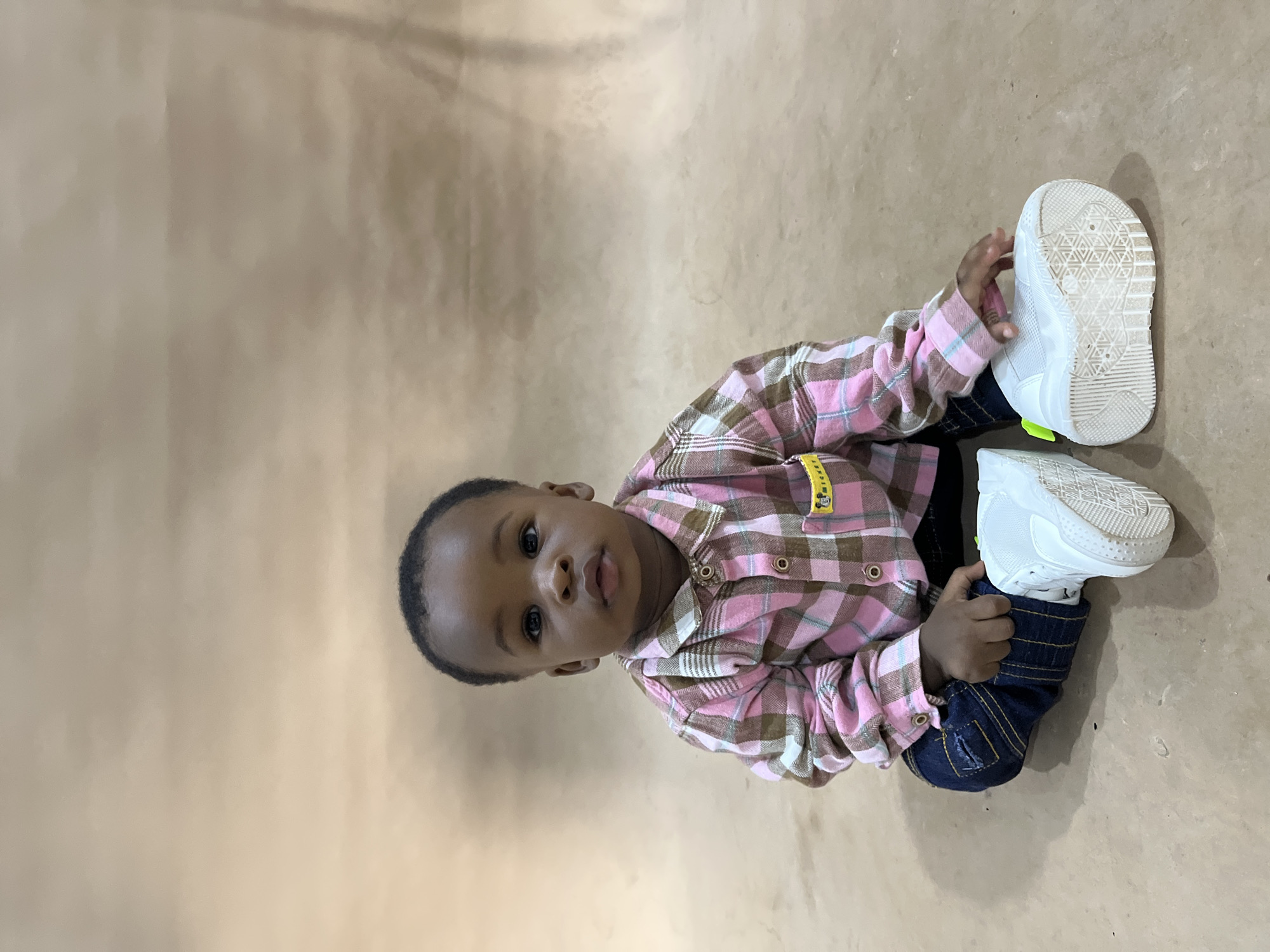 A young Black child, likely less than two years old, is sitting on a light brown, textured surface that appears to be a floor or backdrop. The child is wearing a pink and brown plaid long-sleeved shirt with buttons down the front and a yellow tag with a cartoon character on the left breast pocket. Dark blue denim jeans are visible at the waist. The child is wearing white sneakers with neon yellow accents. The child is looking directly at the camera with a neutral expression. The background is a plain, light brown, slightly mottled surface, suggestive of a studio setting. There are no visible landmarks or elements that would place the image specifically in Gura Top, Nigeria, or any other city. The lighting appears to be soft and even, likely from studio lights.