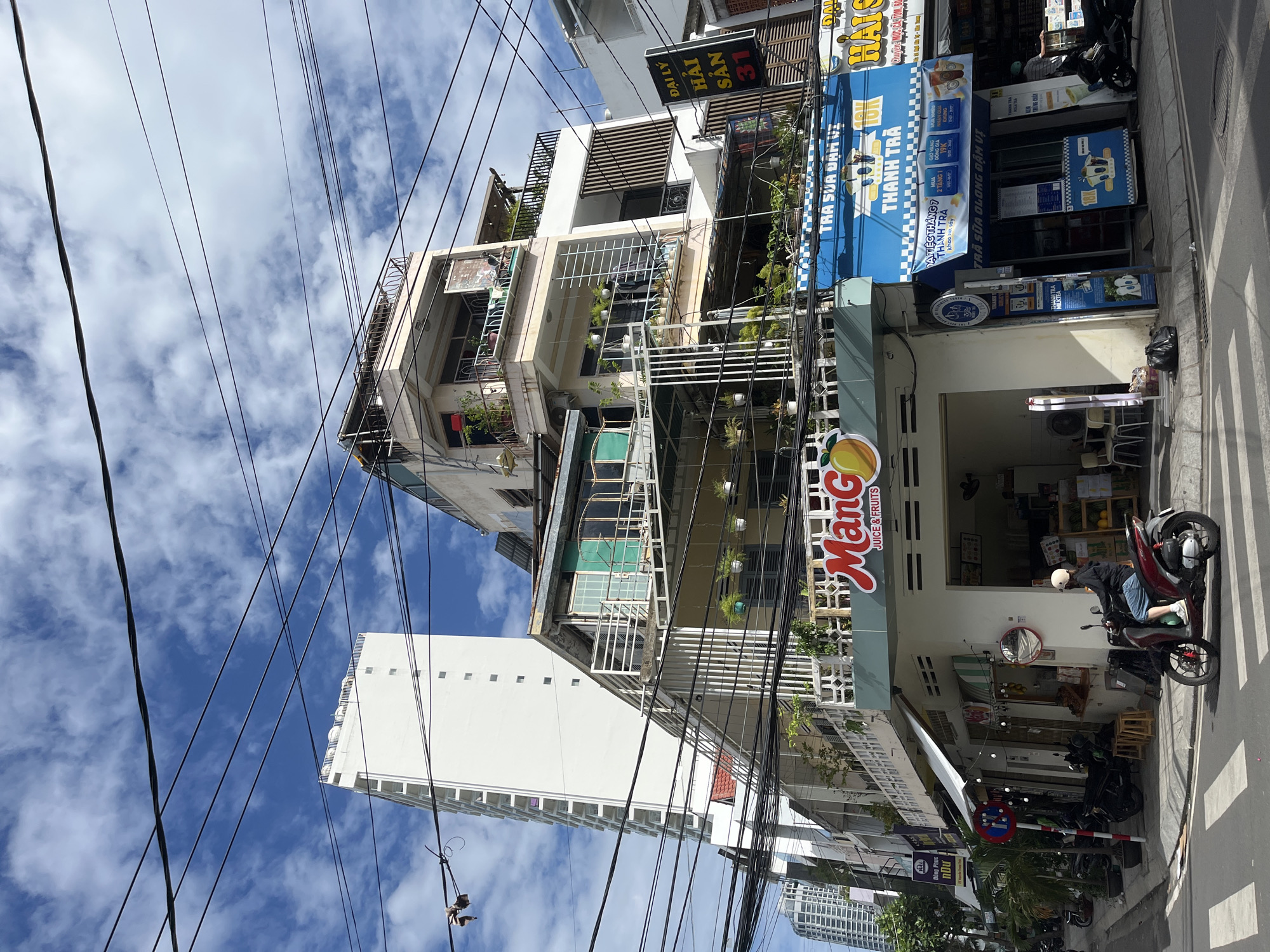 The image captures a street scene in Nha Trang, Vietnam, characterized by a dense network of overhead power lines. In the foreground, a woman on a red scooter is crossing a pedestrian crosswalk. To the right, a storefront with blue awnings displays Vietnamese text, likely advertising bubble tea, with the prominent words "THANH TRA" and "TRA SUA" visible. Further to the right, another storefront displays "ĐẠI LÝ HẢI SẢN 31," indicating a seafood distributor. Behind these street-level businesses, multi-story buildings with balconies and greenery are stacked, and a tall, white modern building rises in the background. The sky is blue with scattered clouds.