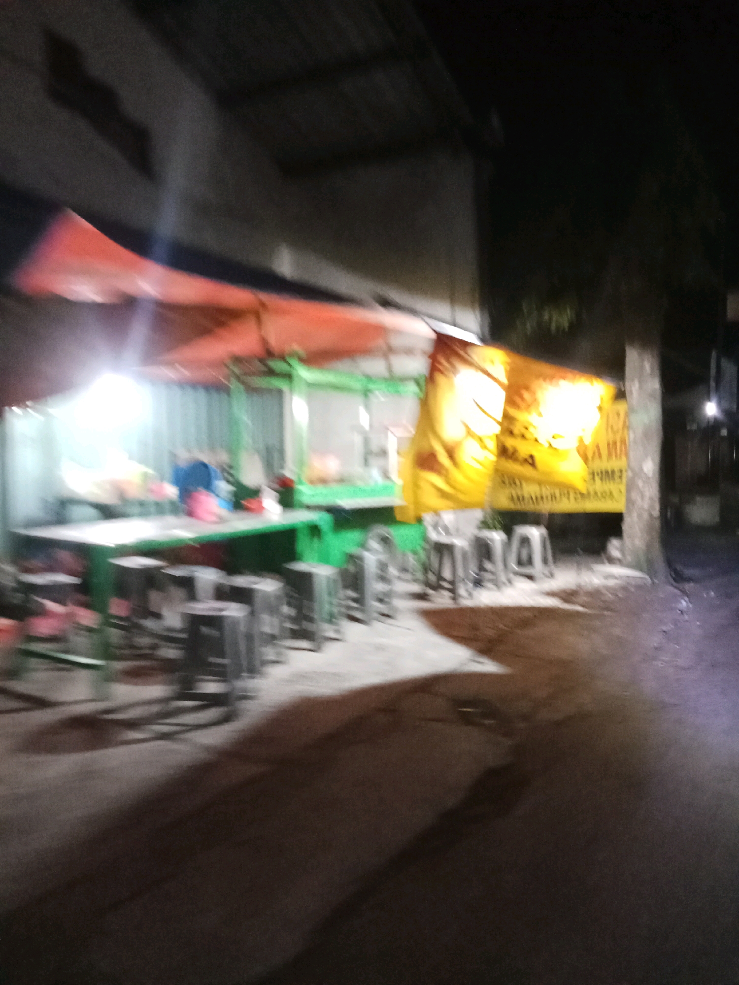 The image depicts an outdoor food stall at night in Kepanjen, Indonesia. The stall features a green table with various items on it, including colorful containers and a blue pitcher. Several stools are arranged around the table. A bright orange awning provides cover, and a large yellow banner with Indonesian text hangs to the right of the stall. The background shows a building and a street, with a tree on the right side. The scene is dimly lit, with artificial lights illuminating the stall area. The overall impression is that of a typical roadside food vendor operating in the evening.