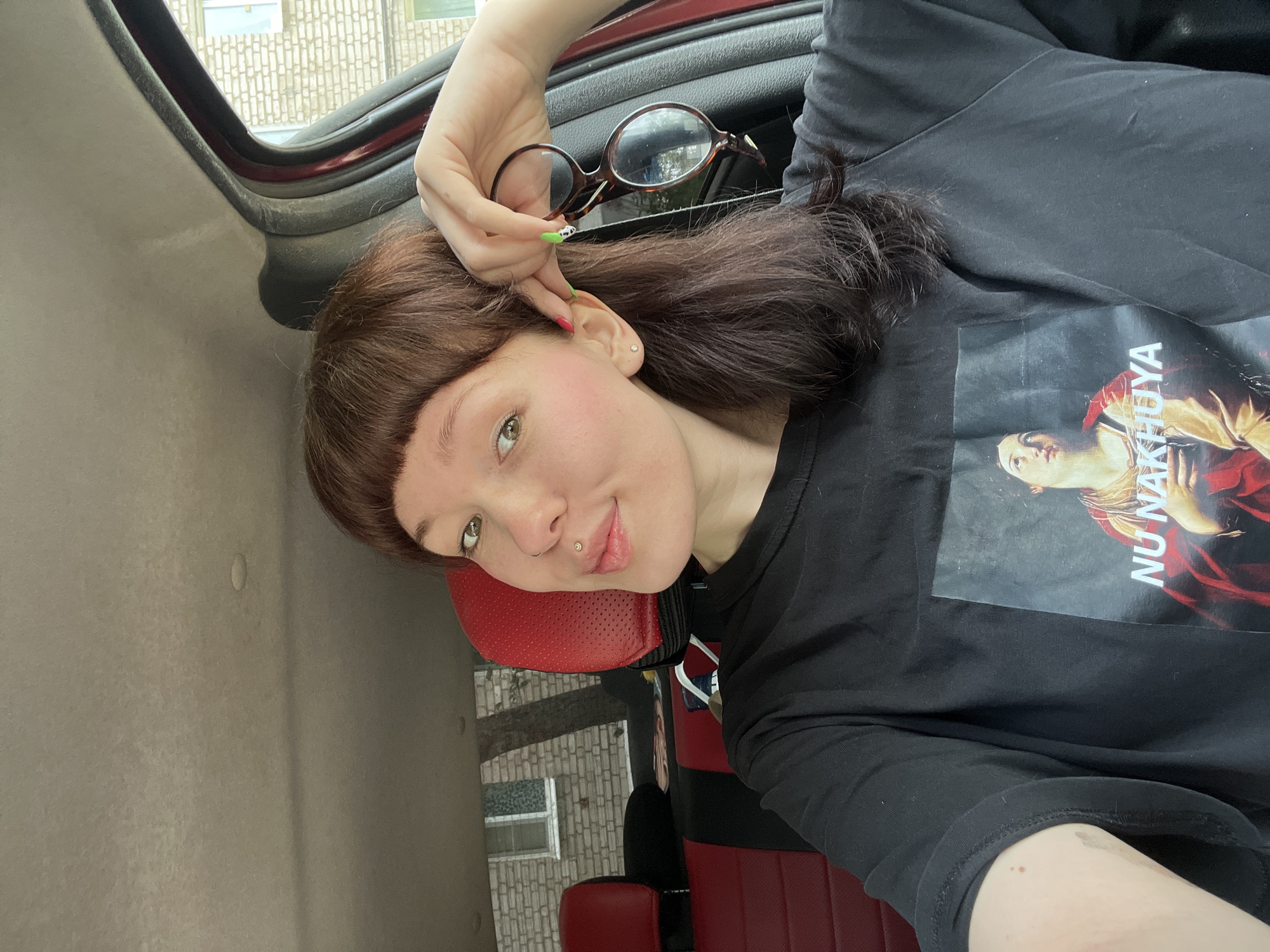 A fair-skinned woman with short bangs, wearing a black t-shirt, reclines in the back of a car. She holds a pair of eyeglasses in her right hand. The car's interior features red seating. Visible through the car window is a brick building typical of urban architecture, suggesting a setting similar to Chaikovsky, Russia. The woman has a lip piercing and an earring in her left ear. Her fingernails are painted with colorful designs.