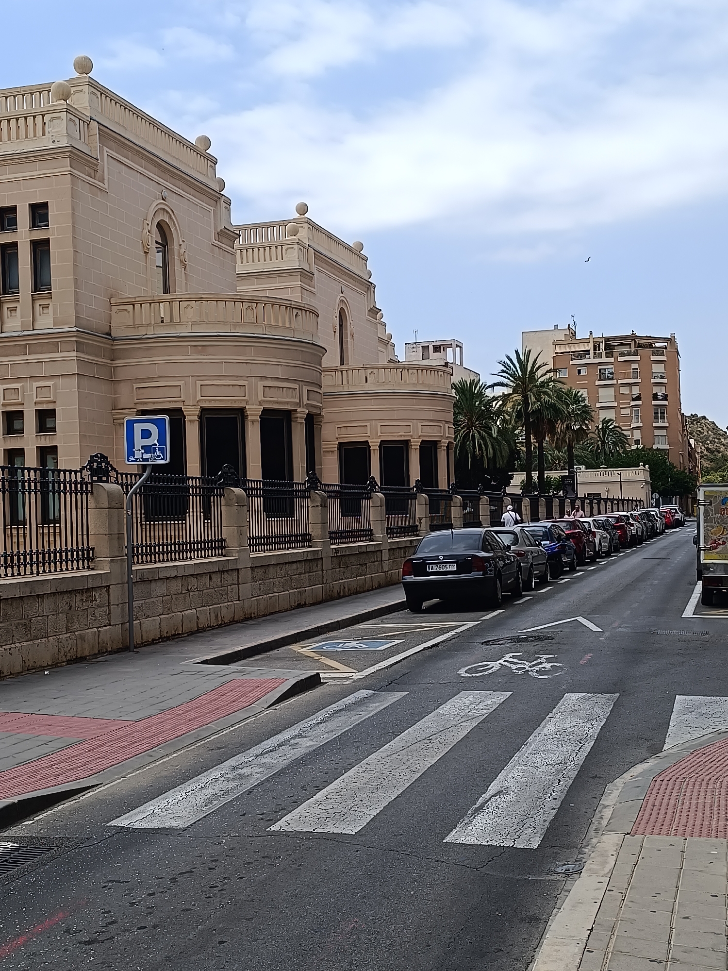 The image depicts a street scene in an urban environment, likely in a Mediterranean climate given the presence of palm trees. On the left side of the street, a large, ornate building with a light-colored stone facade and multiple arched windows and balconies dominates the foreground. A black wrought-iron fence with stone pillars separates the building from the sidewalk. A blue and white sign indicating "P" for parking with an accessible symbol is mounted on a pole in front of the fence.
The street itself is paved and lined with parked cars. A pedestrian crossing with white stripes is visible in the lower part of the image. A bicycle symbol is painted on the road near the pedestrian crossing. A raised red textured tactile paving is present on the sidewalk next to the road, likely for visually impaired pedestrians.
Several cars are parked along the right side of the street, forming a queue. These vehicles are of various colors and models. In the background, taller buildings, including a multi-story brown apartment building with numerous balconies, are visible. The sky is partly cloudy. On the far right, a portion of a truck with a colorful advertisement on its side is visible.
There are a few people visible in the image, including a person walking on the sidewalk and several individuals standing or walking near the parked cars. The overall impression is a typical urban street scene with architecture, traffic, and pedestrian infrastructure.