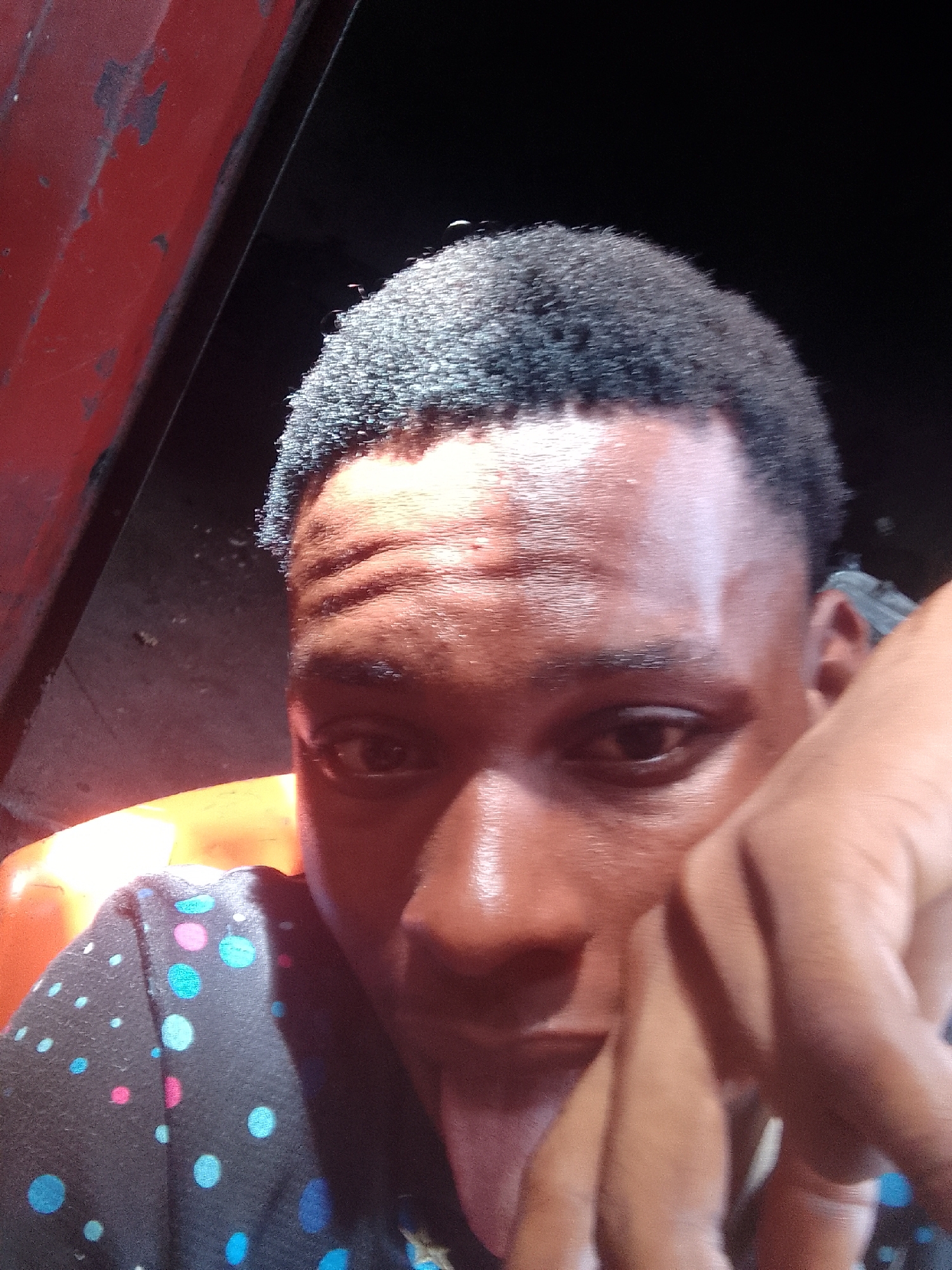 A close-up selfie of a young, dark-skinned man with short, dark hair. He is wearing a dark shirt with light blue and pink polka dots. He is sticking his tongue out slightly. His face appears sweaty, with visible forehead lines. His right hand is partially covering his face. The background is mostly dark, with a portion of a red, possibly metallic object visible on the upper left. Given the request for location awareness and the general skin tone of the subject, it could be a street in Onitsha, Nigeria but there's no visible sign that can identify the location.