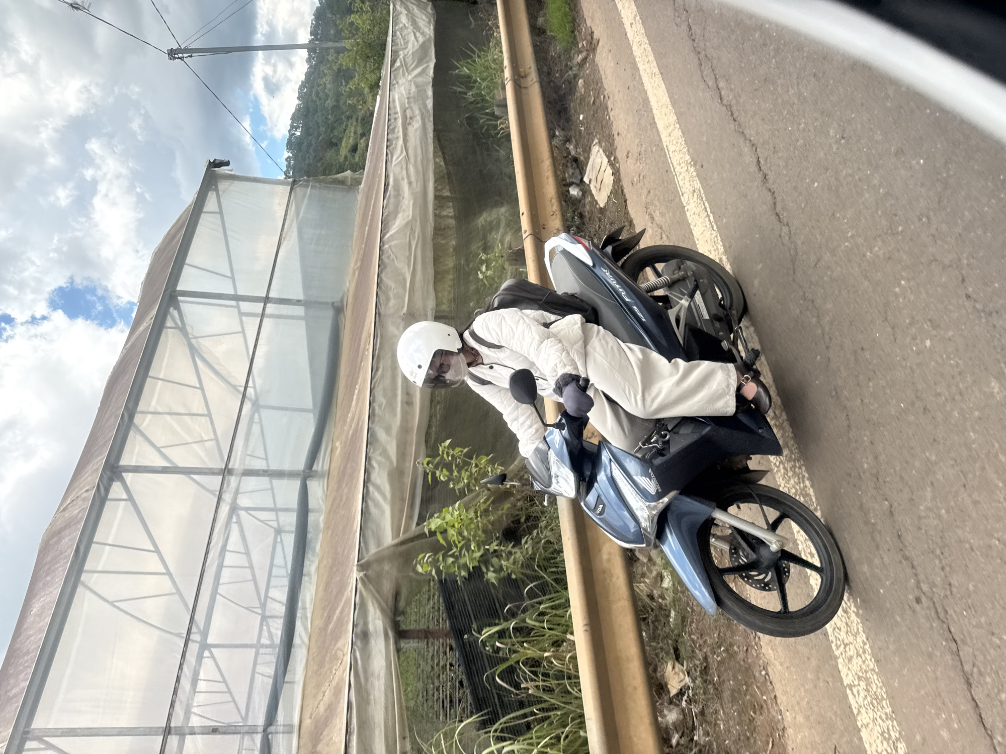A person rides a dark blue Honda motorcycle along a paved road in Đạ Cháy, Vietnam. The rider wears a white helmet, sunglasses, a white quilted jacket, light-colored pants, and dark gloves. A black backpack is visible on their back. The motorcycle has a silver accent and "125 FI" text on the side. The road has a white line marking and appears cracked in places. To the left of the road is a brown guardrail and a grassy embankment. Behind the embankment is a greenhouse structure covered with translucent plastic sheeting, supported by a metal frame. The greenhouse appears to house rows of dark green plants. In the background, a forested hillside is visible under a partly cloudy sky.