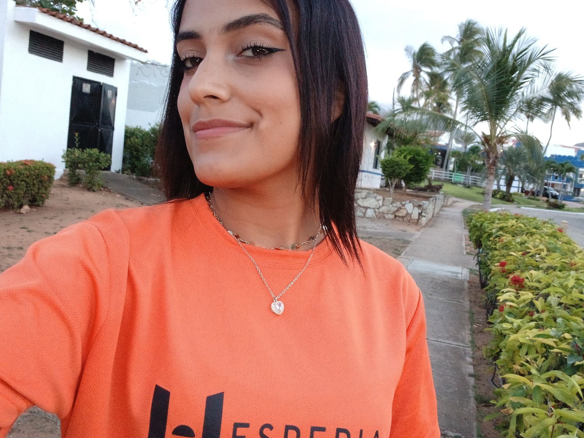 A woman stands outside in Playa El Agua, Venezuela, wearing an orange t-shirt and a heart pendant necklace. She has dark hair and eyeliner. Behind her is a street with foliage and buildings. A palm tree is visible in the distance.