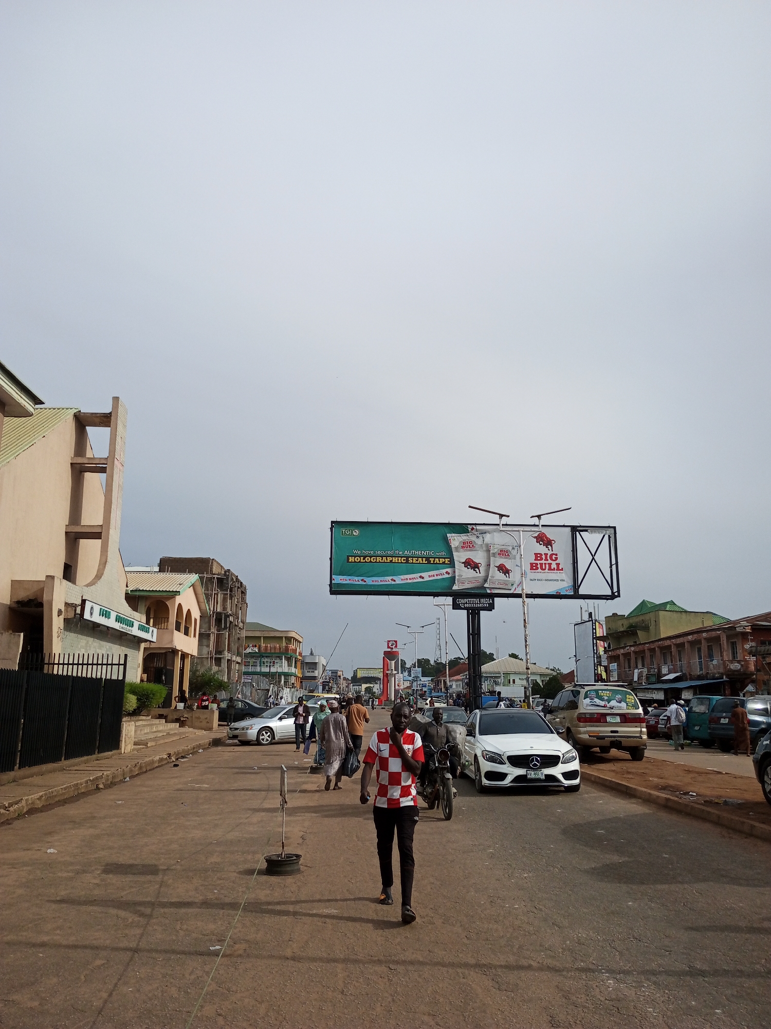 The image shows a street scene in Jos, Nigeria. A man in a red and white checkered shirt and black pants walks towards the camera on a dusty road. Several cars, including a white Mercedes-Benz, are on the road. People are walking on the sidewalks and crossing the street. A large billboard advertising "BIG BULL" holographic seal tape is visible in the background. Buildings on both sides of the street include a light-colored building with a green roof and an orange building with arched windows. The sky is overcast.
