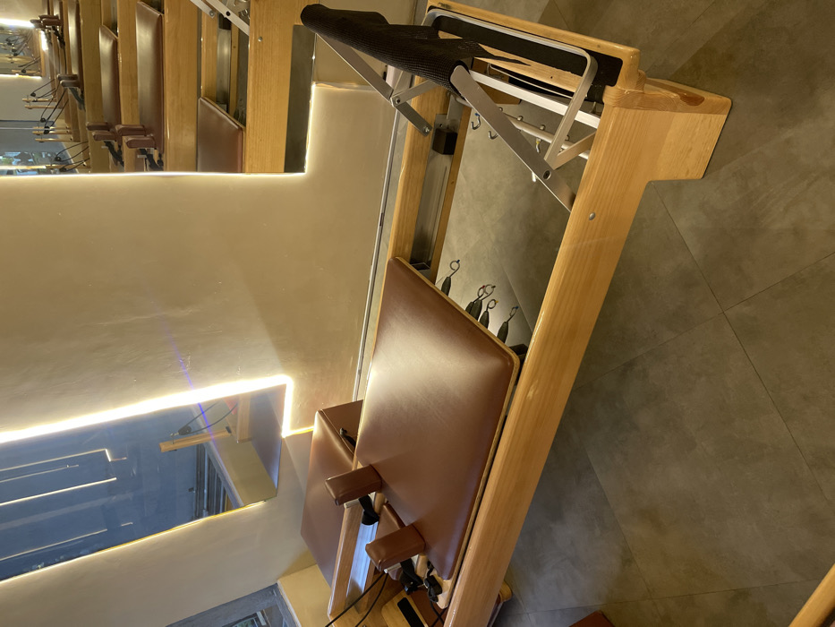 The image displays a brightly lit, unoccupied Pilates studio in Mexico City, Mexico. In the foreground, a light-toned wooden Pilates reformer with brown padded seating, shoulder rests, and a black fabric foot bar is positioned on dark grey tiled flooring. To the left, a large window reflects the studio's interior, including an an L-shaped wall light fixture that emits a warm glow. The background, visible through reflections, reveals multiple identical Pilates reformers arranged in rows, indicative of a professional fitness facility. No individuals, actions, or interactions are depicted.