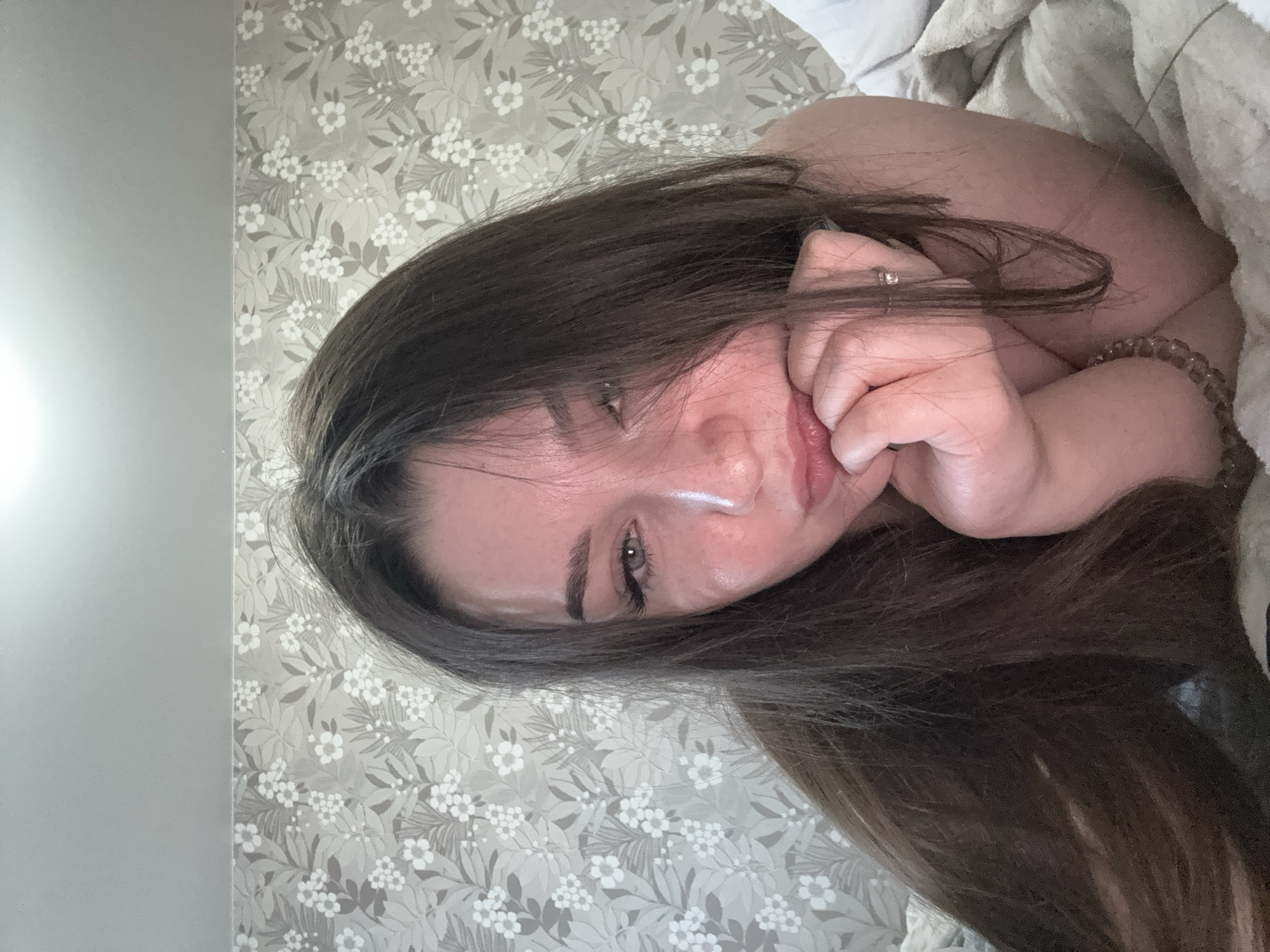 A young woman with long, dark hair is lying on a bed, looking at the camera. Her right hand is resting on her chin, and her left hand is partially covering her mouth. Her eyes are open, and she appears to be looking directly at the camera. The woman is wearing what appears to be a light-colored top, and her shoulders are visible. She has a ring on her left ring finger and a beaded bracelet on her right wrist. The background consists of a gray wall with a floral wallpaper pattern. The lighting suggests it is daytime, possibly in the morning or afternoon. The overall mood is relaxed and intimate. No visible text is present in the image.