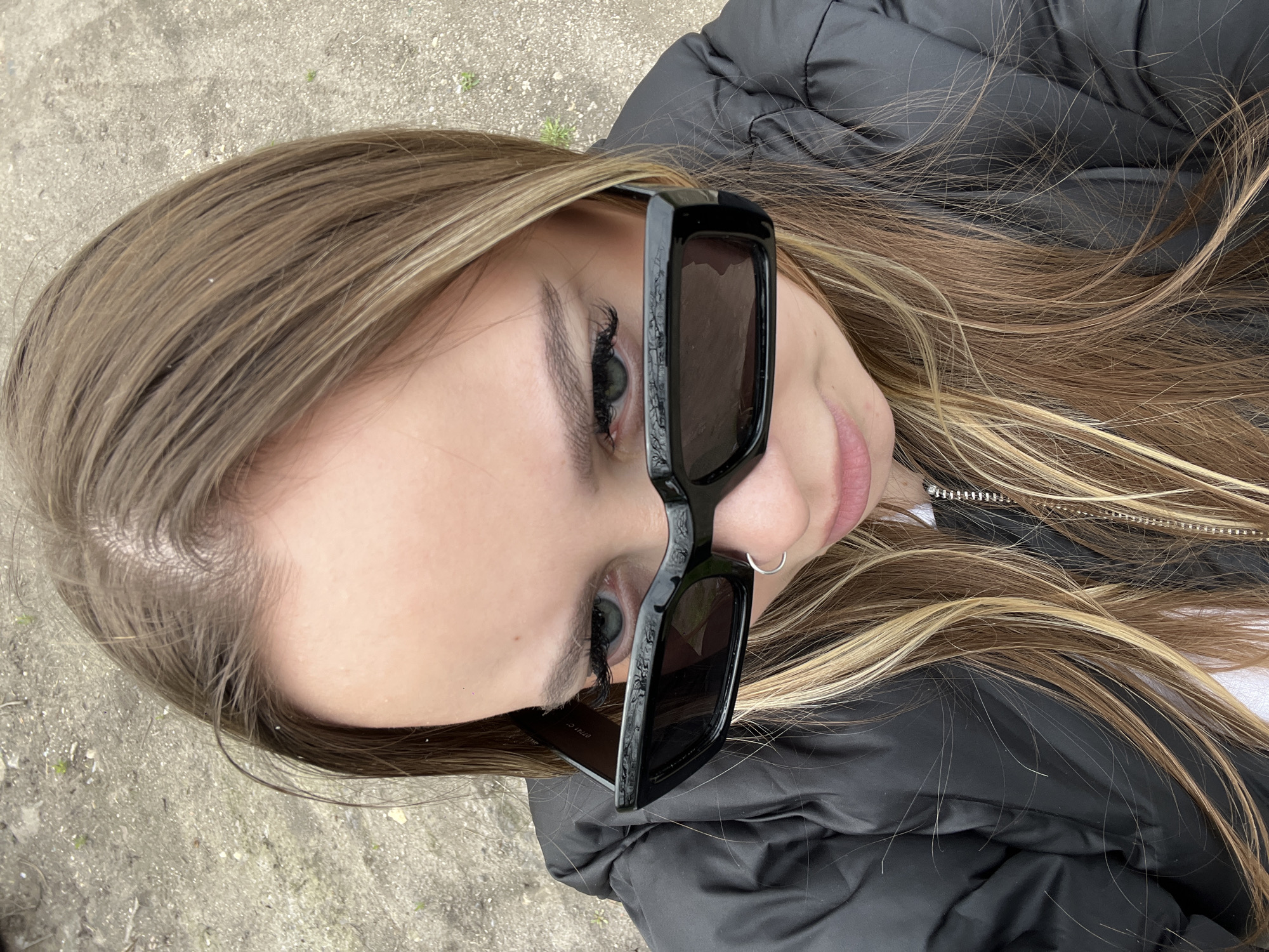 A young woman is pictured from a close-up, slightly high-angle perspective. She is wearing dark, rectangular sunglasses and a black puffer jacket. She has long, light brown hair with blonde highlights. An earring is visible in her left ear. The background shows a patch of concrete, suggesting an urban setting. There are no indications of location in the city of Port-Katon, Russia.