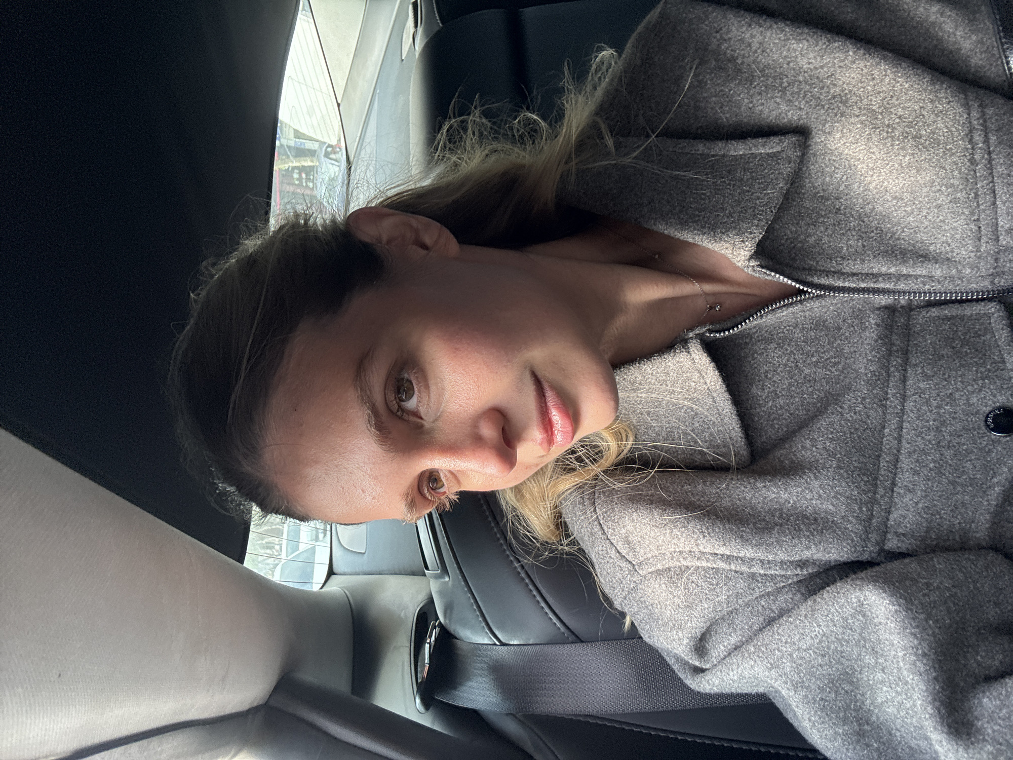 A young woman is captured in a close-up, horizontally oriented shot, sitting inside a car. She has light brown hair with blonde highlights, brown eyes, and a natural complexion, looking directly at the camera with a soft, engaging expression and a subtle hint of a smile. She is wearing a grey, textured, possibly wool or felt, coat with a zipper. A delicate silver chain with a small, clear pendant is visible around her neck, peeking out from the partially unzipped coat.
The setting appears to be the back seat of a vehicle, featuring black leather upholstery and a seatbelt visible across the lower portion of the frame. Bright natural daylight enters from the car window on the left, casting illumination on her face and hair. The background visible through the window is blurry but suggests an urban environment with structural elements, consistent with a city like Tbilisi, Georgia. The activity taking place is a posed photograph, likely a selfie. No visible text is present in the image.