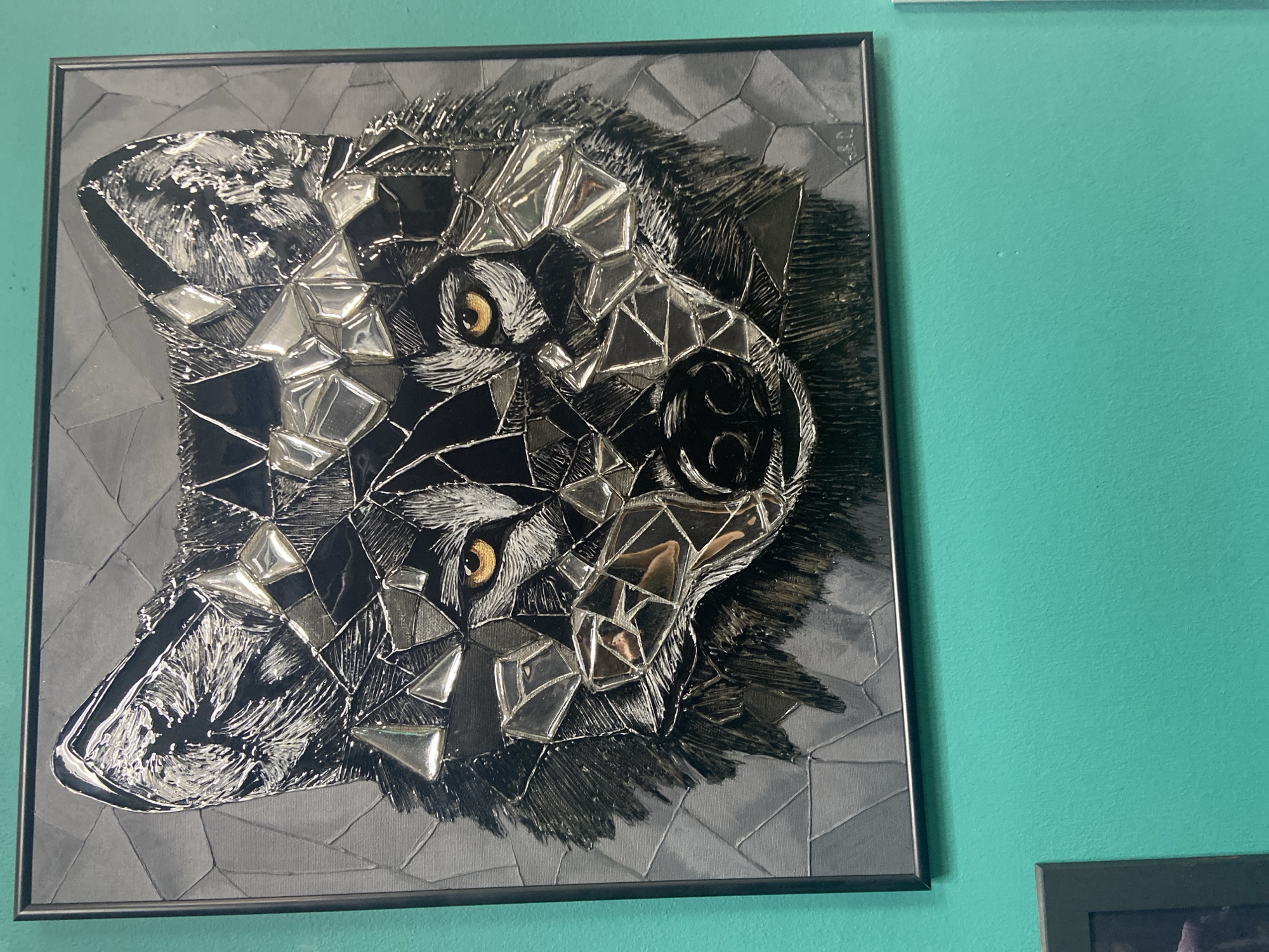 This is a photograph of a framed mosaic artwork depicting a wolf's head, rendered in black and silver, with bright yellow eyes. The artwork is hung on a turquoise wall. The mosaic is composed of fragmented pieces, many of which appear to be reflective, giving the impression of broken glass or mirrors. The background of the artwork is a textured gray, with geometric patterns. The wolf's fur is depicted with fine lines and shading, and the muzzle and nose are detailed. A signature or initials are visible in the bottom right corner of the artwork. The overall impression is one of a modern, artistic representation of a wolf. The location is Yegorlykskaya, Russia.