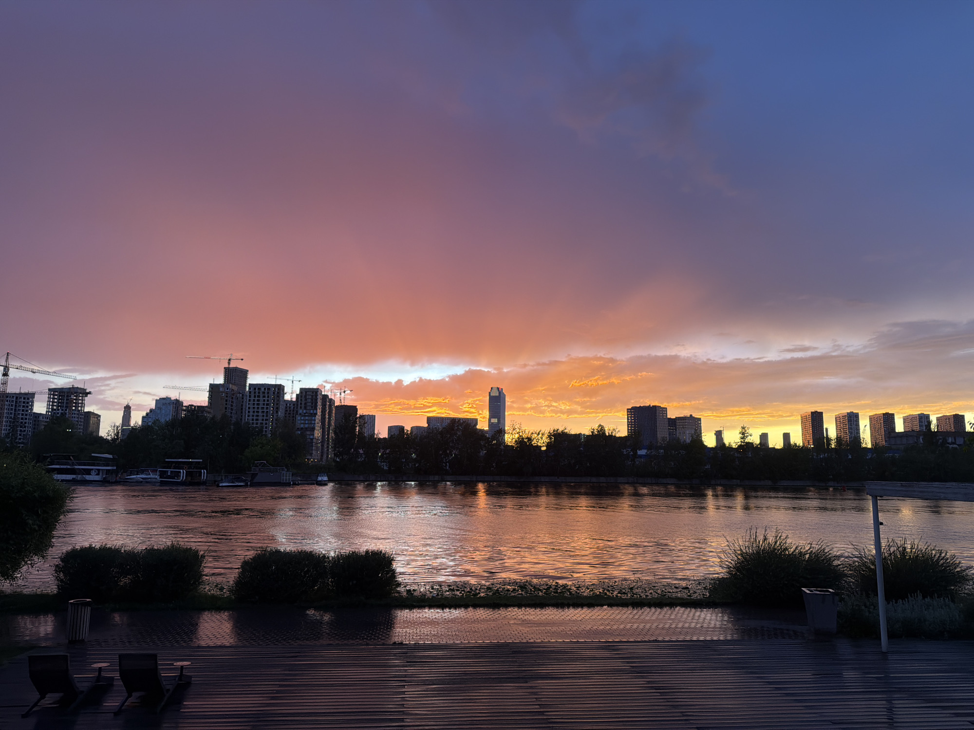 A serene, wide-angle view captures a tranquil river reflecting the dramatic hues of a sunset over a city skyline. The sky is a gradient of deep blues at the top, transitioning through soft purples and vibrant oranges and yellows near the horizon, where the sun has just set. Wispy clouds are illuminated by the fading light, adding texture to the celestial canvas.
Along the riverbank, a dense line of trees forms a dark silhouette against the vibrant sky. Behind the trees, a modern cityscape rises, featuring numerous residential buildings and construction cranes, indicating ongoing development. A prominent tall skyscraper with a distinct shape stands out in the center of the skyline. The river itself is calm, with gentle ripples disturbing its surface and mirroring the colors of the sky. A few boats and houseboats are moored along the bank, suggesting a leisurely riverside environment.
In the foreground, a wooden deck provides a vantage point overlooking the scene. Two empty lounge chairs and small side tables are positioned on the deck, hinting at a place for relaxation and observation. The deck's surface appears wet, possibly from recent rain or dew, and reflects the subdued light. Two large, dark green bushes are planted along the edge of the deck, closer to the river. A trash receptacle is visible near the bushes.
The overall mood is peaceful and contemplative, evoking a sense of calm as day transitions into night. The time of day is clearly sunset, with the sky's colors being the most prominent indicator. The location is identified as Moscow, Russia, which aligns with the architectural style of the buildings and the presence of a significant river in a developed urban area. No visible text is present in the image.