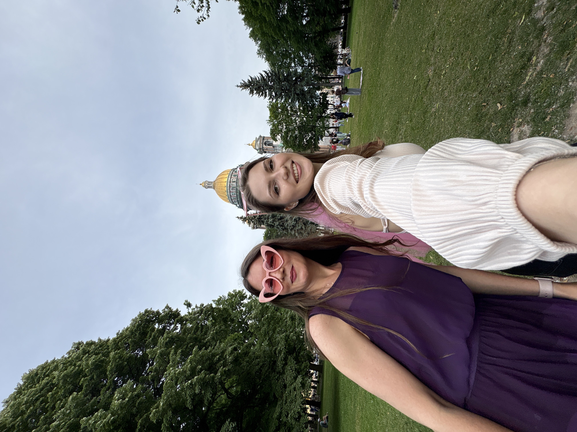 Two women are taking a selfie in a park in Saint Petersburg, Russia. In the background, Saint Isaac's Cathedral is visible with its golden dome. The woman on the left is wearing a purple dress and pink heart-shaped glasses. The woman on the right is wearing a white, puffed-sleeve top. Both women are smiling. The park is green with trees and some people are scattered in the background. The sky is overcast.