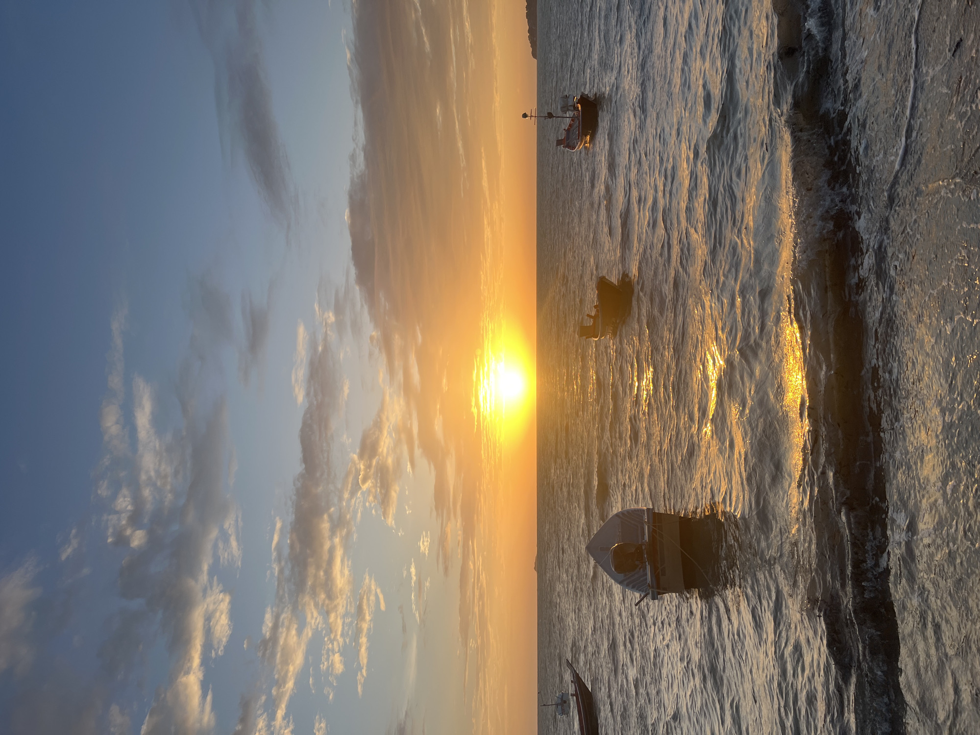 This image captures a serene sunset over the ocean in Ko Pha-ngan District, Thailand. The sun, a brilliant orb, is low on the horizon, casting a warm, golden glow across the water and illuminating a scattering of white and grey clouds in the sky. Several small boats are visible on the calm sea, some anchored near the shore, while others are further out. The water is gently rippling, reflecting the vibrant colors of the sunset. The foreground shows a shoreline with small waves lapping at the edge. The overall atmosphere is one of tranquility and natural beauty.
