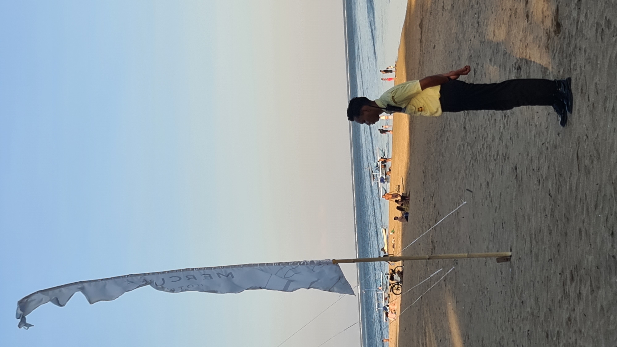 This media file captures a scene at a beach in Denpasar, Indonesia. The sun appears to be setting, casting a warm glow on the sandy shore and the calm ocean. A person in a yellow shirt and black pants is standing on the beach, their body oriented horizontally as if performing a plank exercise. Their hands are clasped behind their back, and their head is bowed. In the foreground, a white flag with some text is unfurled, attached to a bamboo pole. The flag is tattered at the edges. In the background, several other people are visible on the beach, some relaxing on lounge chairs or walking near the water's edge. The overall atmosphere seems peaceful and relaxed, indicative of a typical beach day. The visible text on the flag appears to be "WEBCON HOTEL".