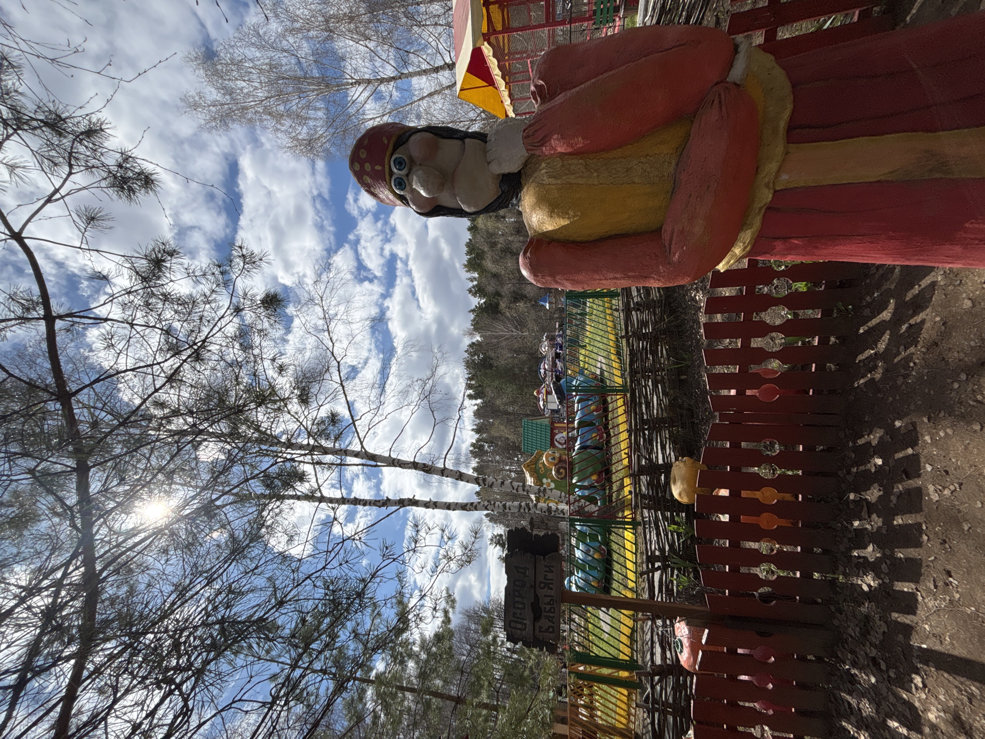 The image shows a statue of a woman in Chaikovsky, Russia. The statue is wearing a red hat with white polka dots, a yellow blouse, and a red skirt. The statue is standing next to a red fence. In the background, there is a ride for children and some trees. The sky is blue with white clouds.