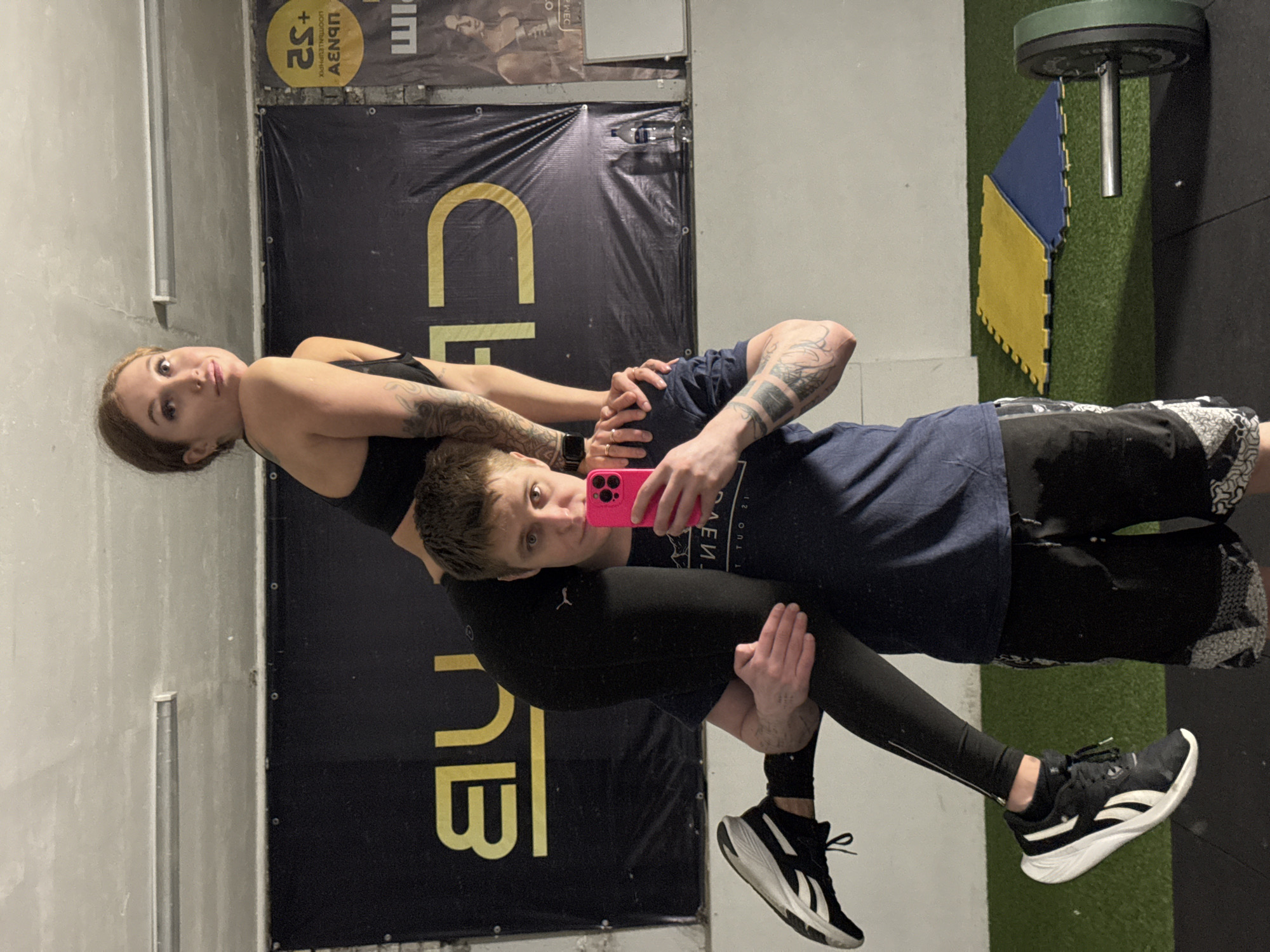 An indoor scene, likely a gym or fitness studio, features two individuals performing a partner acrobatic pose. The male, positioned on the bottom, supports the female upside down on his shoulders. He wears a dark blue t-shirt, dark shorts over black leggings, and black and white Reebok sneakers. He has tattoos on his arms and holds a bright pink smartphone, taking a selfie while looking at the screen. The female, atop the male, wears a black sports bra and black leggings, also displaying arm tattoos. She looks directly at the camera.
The background consists of a light grey wall on the left and a large black banner with partially visible yellow text, which appears to spell "CLUB" or a similar fitness-related word. Above this banner, another, smaller banner displays fragmented, mirrored, and upside-down Cyrillic text that includes "ЦЕНЫ" (prices) and possibly "+S2" or "+52". The floor is covered with green artificial turf, accented by interlocking blue and yellow puzzle mats. A weight plate is visible on a rack in the upper right corner of the frame. The overall impression is one of an active and athletic environment.
