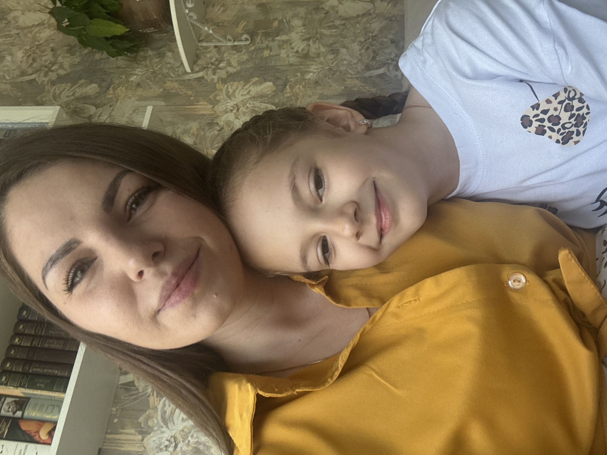 A selfie shows a woman and a young girl. The woman has medium-length brown hair and is wearing a yellow shirt. The girl has her hair in braids and is wearing a white shirt with a leopard print heart on it. The girl is resting her head on the woman's chest. There are books and a plant visible in the background. The location is Раздолье, Russia.