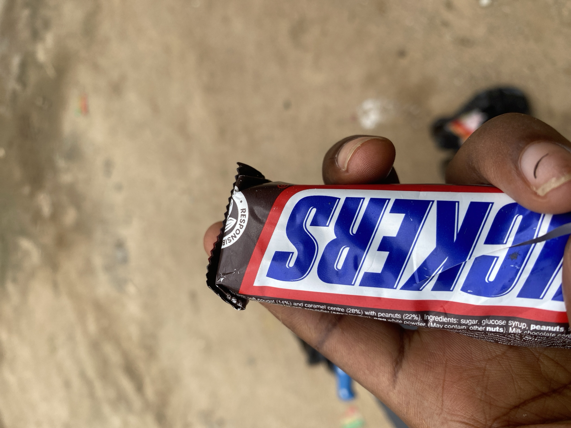 A close-up photograph features a dark-skinned hand holding a partially opened Snickers candy bar. The wrapper prominently displays the blue "SNICKERS" logo on a red and white background. Visible text on the wrapper includes details about the nougat (14%), caramel centre (28%), and peanuts (22%), along with "Ingredients: sugar, glucose syrup, peanuts." The top, folded-back portion of the brown wrapper shows the word "RESPONSE." The hand's thumb and index finger are visible, with natural fingernails. The background is a heavily out-of-focus, light brown, unpaved ground, appearing as dirt or sand. Distant, blurry shapes include a faint blue object on the mid-left, a small, colorful (red and green) object in the far center, and a dark object with orange/red accents on the mid-right. The scene is located in Kubwa, Nigeria.
