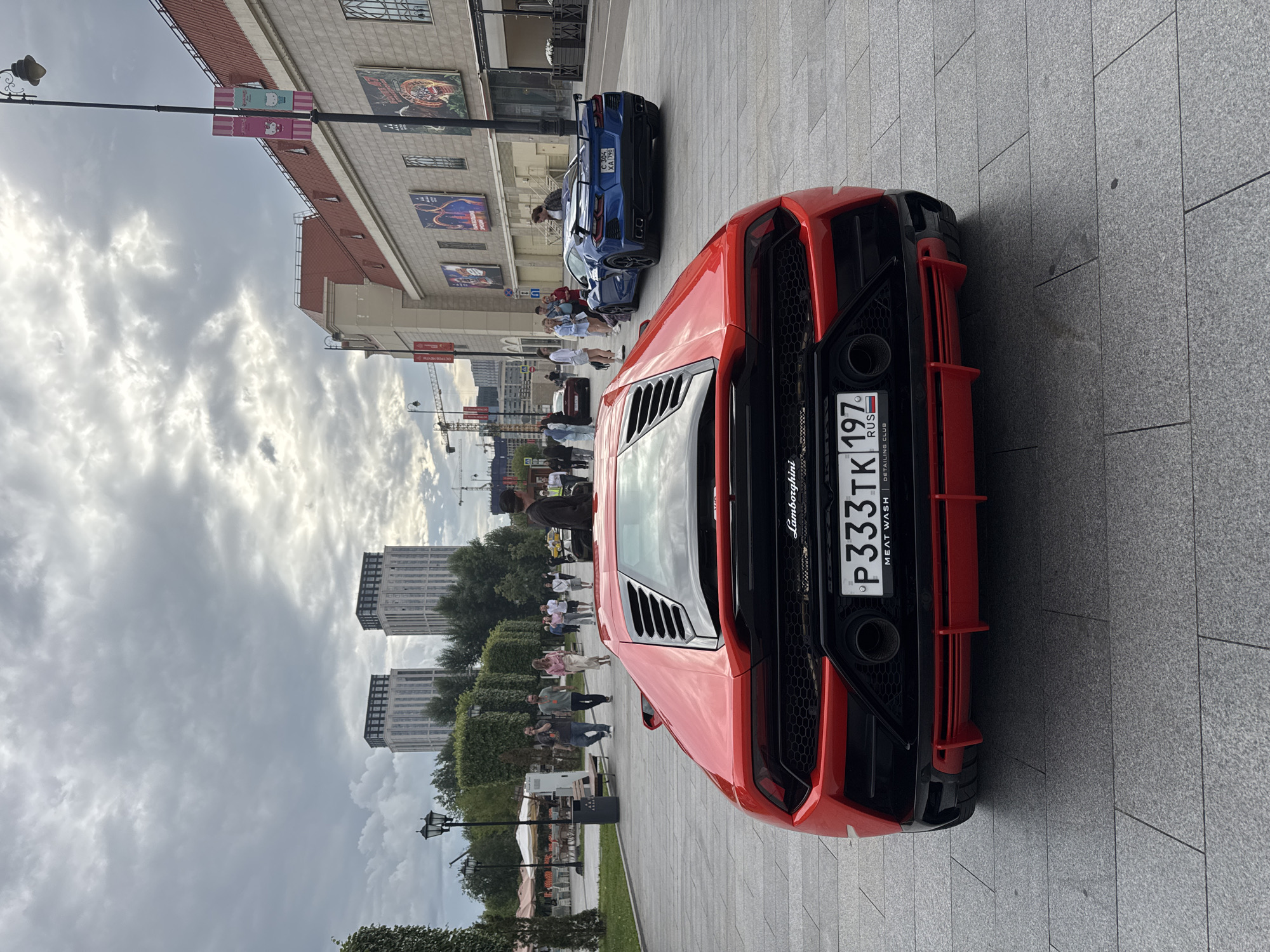 This image depicts an urban scene with a focus on two sports cars. In the foreground, a vibrant red Lamborghini is prominently displayed, viewed from the rear. Its sleek design, complete with aggressive styling and a large exhaust system, is evident. The car features a license plate that reads "P 333TK 197 RUS," and below it, "MEAT WASH DETAILING CLUB."
Behind the red Lamborghini, a blue sports car, possibly a Chevrolet Corvette, is parked. It also has a visible license plate. Several people are gathered around the cars, some standing and observing, while others appear to be walking by. The people are dressed in casual, summery attire, suggesting a mild or warm day.
The setting is a paved plaza or walkway, likely in a city center, given the modern building in the background with large windows and signage. The architecture suggests a commercial or entertainment complex. Further in the background, tall apartment buildings and trees are visible, along with streetlights. The sky is overcast with fluffy clouds, indicating a cloudy day. The overall atmosphere appears relaxed and informal, possibly a car enthusiast gathering or a casual display of luxury vehicles. The image was taken in Moscow, Russia, as indicated by the "RUS" on the license plate and the overall context. The time of day seems to be daytime, given the ambient light.