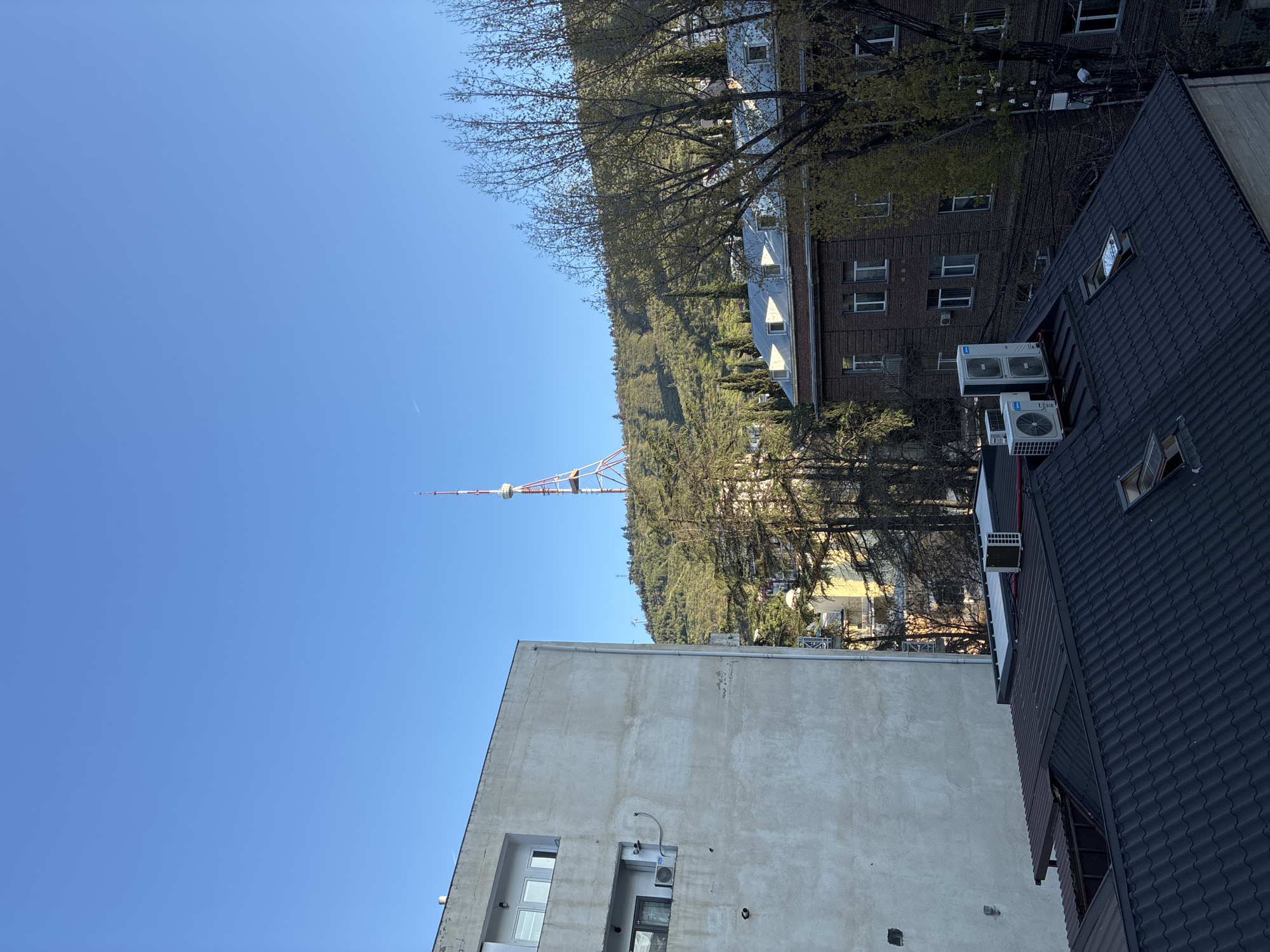 This media file presents an outdoor panoramic view of a cityscape, set against a clear blue sky, with a prominent telecommunications tower marking the horizon. The scene is consistent with Tbilisi, Georgia, featuring its iconic TV tower.
In the immediate foreground, the image captures a dark, possibly tiled or corrugated, rooftop. Adjacent to this roof, a building wall is visible, on which several outdoor air conditioning units are mounted. To the left, another building with a light-colored, stucco or concrete facade is visible, featuring rectangular windows and another air conditioning unit.
Beyond these foreground structures, a verdant hillside rises, densely covered with a mix of deciduous trees, some with bare branches suggesting late winter or early spring, and others already in full green foliage. Several multi-story buildings, varying in architectural style from light-colored facades to brick, are interspersed among the trees on the hillside, indicating residential or commercial development integrated into the natural landscape.
Dominating the distant skyline, atop the forested hill, is the distinct Tbilisi TV Broadcasting Tower. Its reddish-orange, lattice-like structure stands out against the backdrop of a bright, cloudless blue sky. A faint white contrail from an aircraft is visible high in the sky to the left.
The image conveys a sense of a sunny day, likely morning or early afternoon, given the clear conditions and illumination. No people or specific activities are depicted. The composition highlights the blend of urban life with the surrounding natural environment, with the instantly recognizable TV tower serving as a key geographical indicator. No visible text is present within the image.