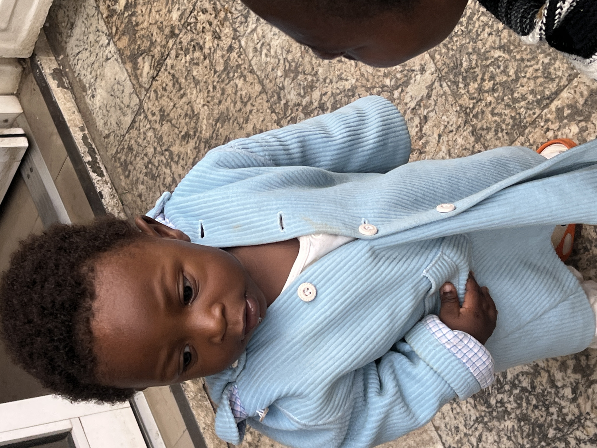An eye-level shot depicts a young child lying on a patterned tiled surface, possibly a floor. The child, with dark hair, dark eyes, and brown skin, is wearing a light blue corduroy coat over a white garment. The coat is partially buttoned. A hand is visible near the child's stomach. A small portion of an adult figure is visible in the top center, while another person is wearing a striped patterned fabric in the top right corner of the image. A window frame is visible in the bottom left corner of the image. Based on the prompt's context, the location is inferred to be Gura Top, Nigeria.