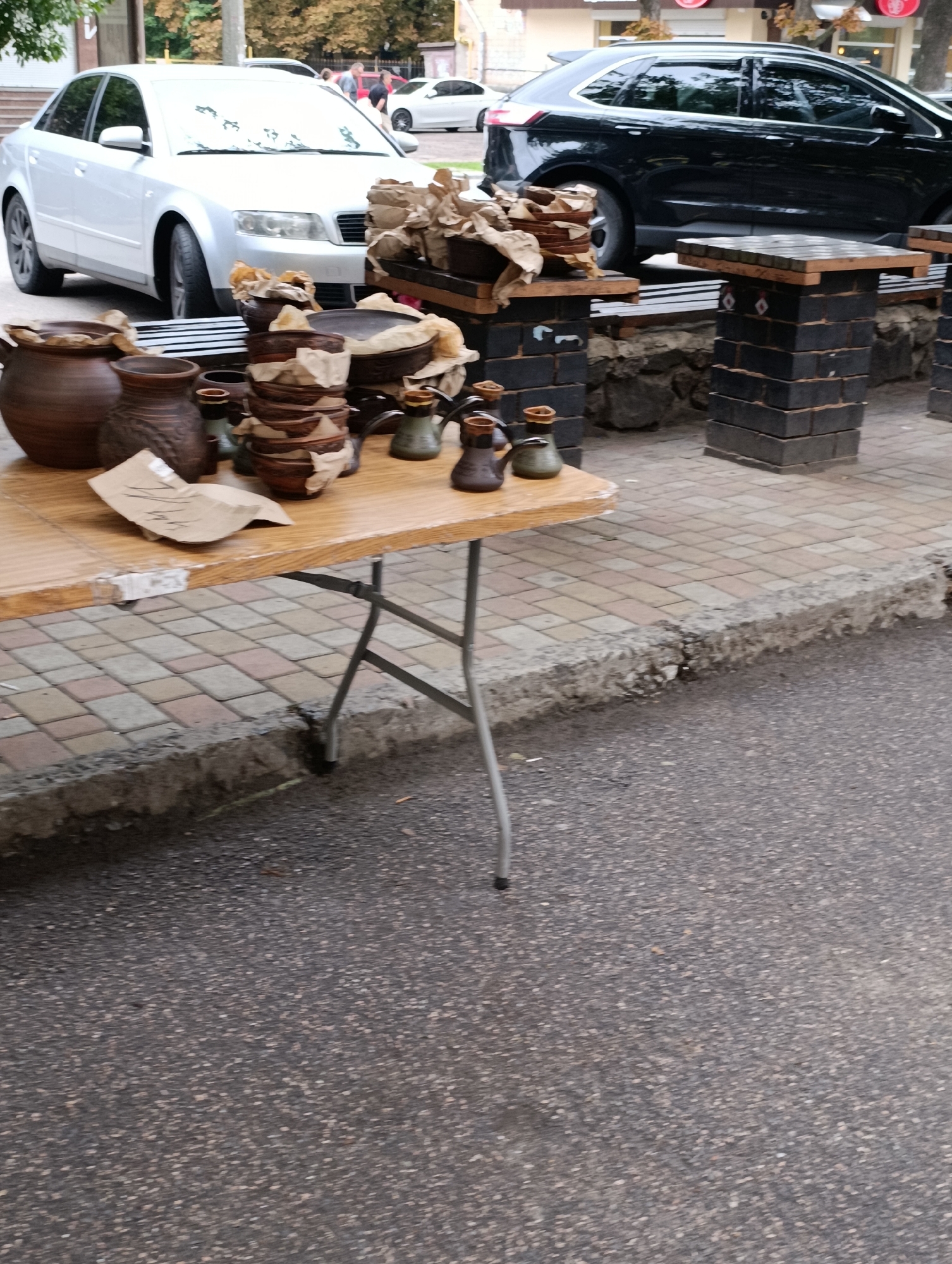 The image shows a street scene in Cherkasy, Ukraine, with a focus on a table displaying various ceramic items. A silver sedan is parked on the left, and a black SUV is parked on the right. The table, made of wood, is set up on a paved sidewalk made of interlocking bricks. On the table, there are numerous clay pots, bowls, and small pitchers, some wrapped in paper. Behind the table, there are two brick pillars supporting wooden surfaces. The ground in front of the sidewalk is asphalt. There are no people clearly visible in the immediate foreground or interacting with the displayed items. The overall scene suggests a street market or an outdoor display of handcrafted goods.