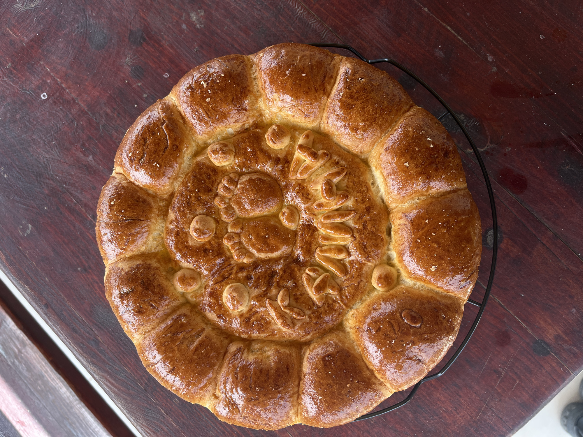 The image shows a loaf of intricately decorated bread on a black wire rack. The bread has a golden-brown, shiny crust. The center is decorated with raised dough, forming shapes reminiscent of animals, leaves, or other symbolic designs. The outer ring is comprised of connected dough balls. The bread sits on a dark-stained wooden surface with visible grain. The picture appears to have been taken indoors in Kostinbrod, Bulgaria, because similar breads are traditional for celebrations in the region.