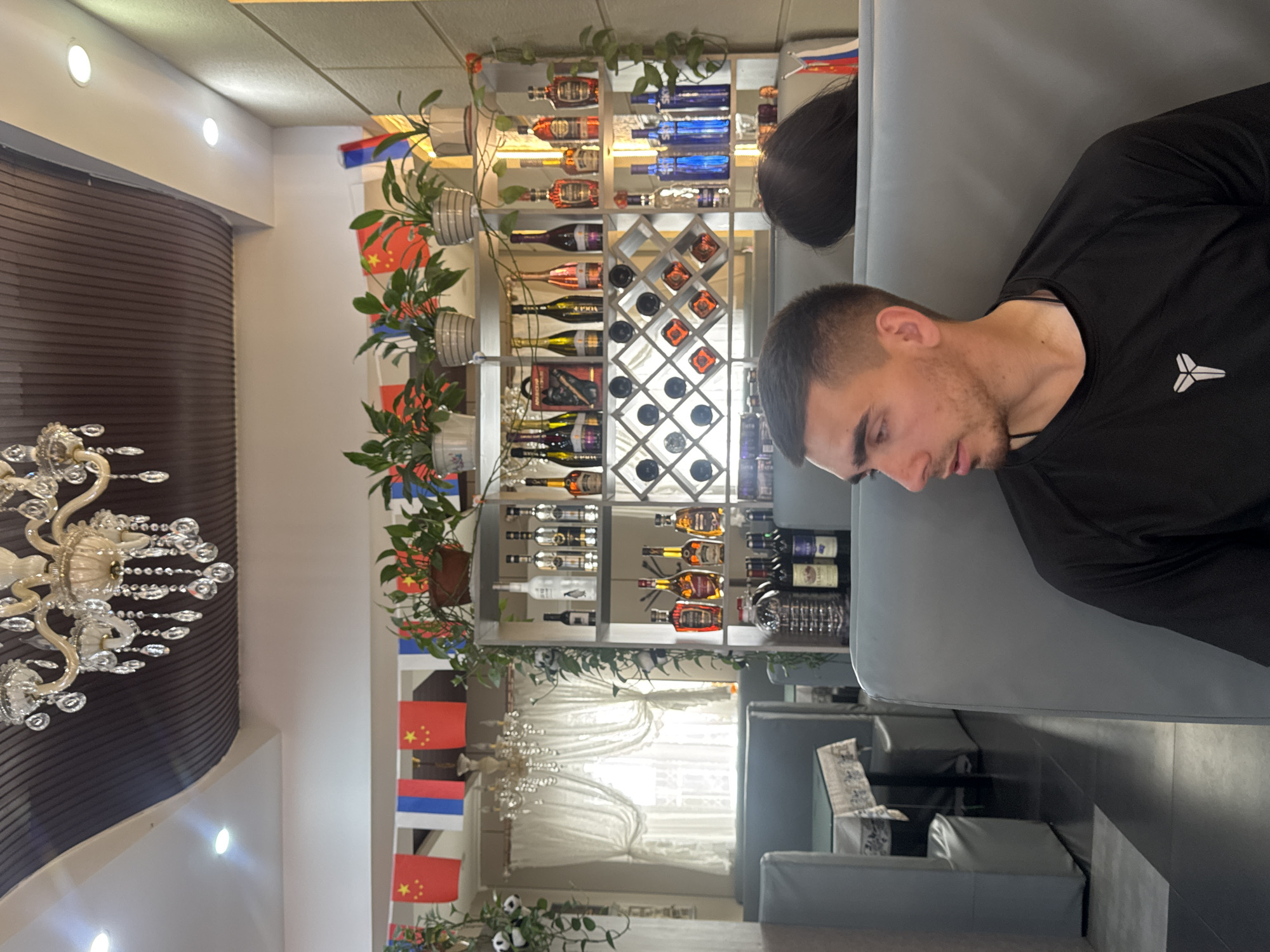 The image depicts an interior scene within a restaurant in Suifenhe, China. A young man with short, dark hair and a light beard is seated on a grey booth, looking to his right. He is wearing a black t-shirt with a white symbol on the left chest. Another person, partially visible and with dark hair, is seated behind him. The restaurant features a decorative chandelier hanging from a dark, ribbed ceiling. Along the back wall, there are shelves stocked with various bottles, including what appear to be spirits and wines. A wine rack with multiple compartments is also visible. In the background, curtains are drawn across a window, and there are flag decorations, including Chinese flags, hanging from the ceiling. Tables with tablecloths are set up in the dining area. The overall environment suggests a casual dining establishment.