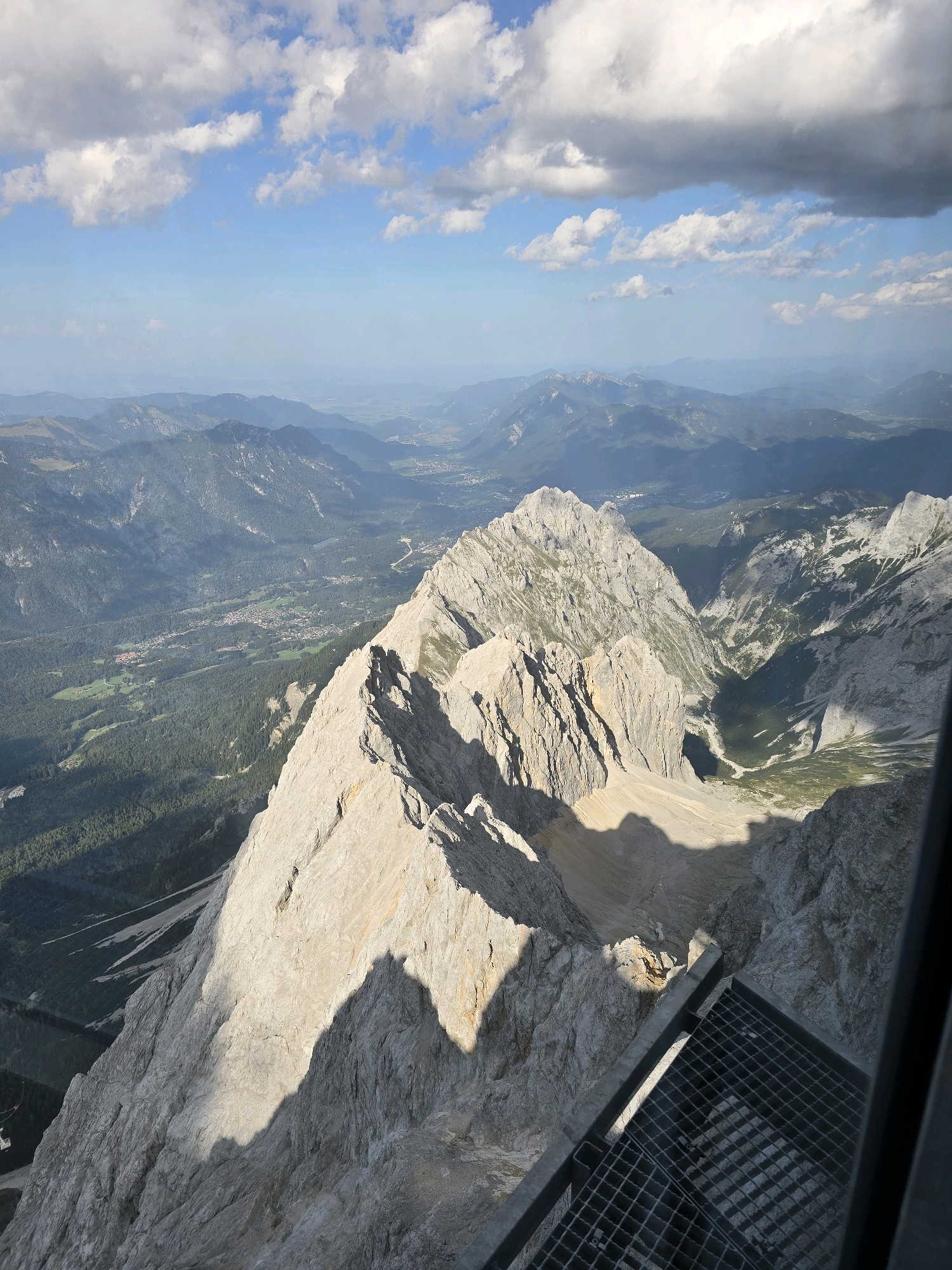 This is a high-angle, close-up aerial shot looking down at the Zugspitze, Germany's highest peak, located near Grainau. The foreground is dominated by the rugged, sunlit, rocky terrain of the mountain summit. A metal grate viewing platform is visible in the lower right corner, with its grid pattern casting shadows on the rocks. In the middle ground, another rocky ridge descends sharply. Further in the distance, a valley unfolds, with dense green forests covering the slopes of lower mountains. A small town or village with scattered buildings and fields is visible nestled within the valley, with a road winding through it. The background features layers of progressively more distant mountain ranges, fading into a hazy blue. The sky above is a bright blue, dotted with white and gray cumulus clouds. The lighting suggests a bright, sunny day.