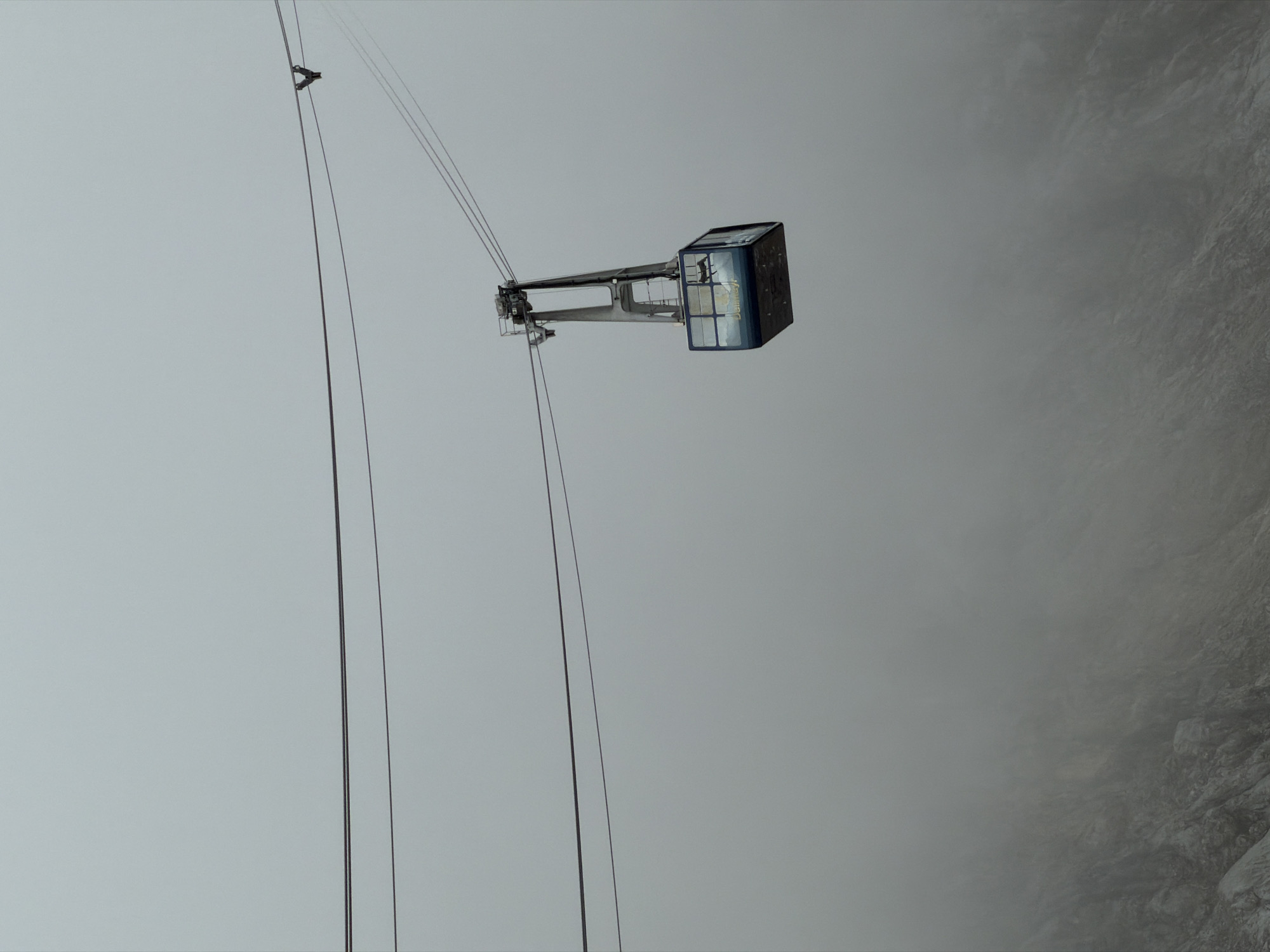A cable car is suspended by cables against a heavily overcast sky. The cable car, which appears to be made of blue glass and metal, is positioned in the center right of the frame. The lower portion of the image shows a hazy, rocky mountain slope obscured by mist. The environment suggests a high-altitude location, likely on Zugspitze mountain, near Garmisch-Partenkirchen, Germany, known for its cable car systems and mountainous terrain. There are no people visible in the frame.