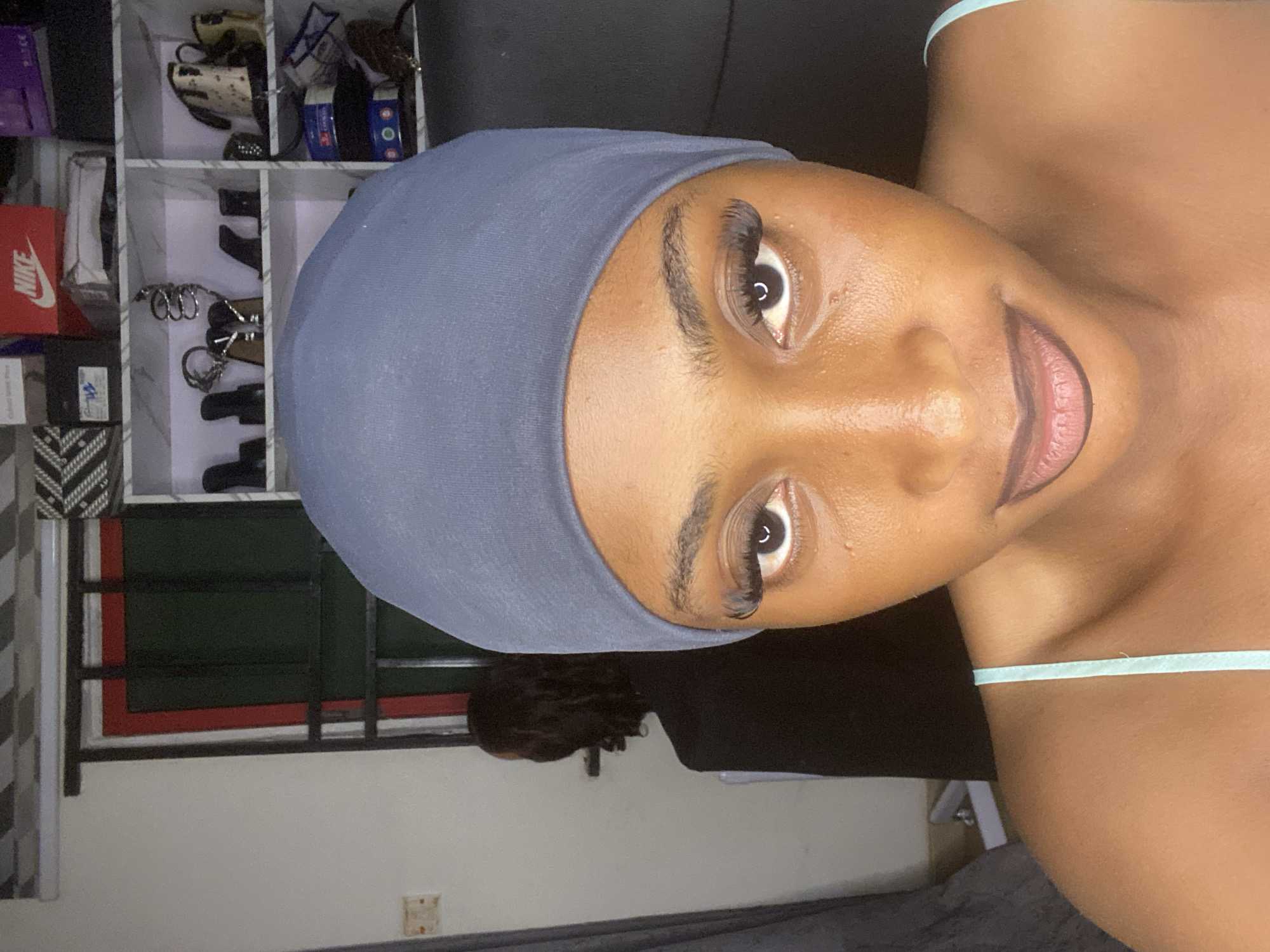 A young Black woman, wearing a blue bonnet and a light blue strap top, is pictured from the chest up, in a close-up selfie. She has on dramatic false eyelashes and dark lipstick. Her skin is smooth and appears to have a dewy finish. In the background, out of focus, is a shelving unit filled with various items, including shoes, boxes, and what appears to be a kettle. To the left of the shelves, a window with green and red accents is visible. A person's dark hair is visible to the left of the woman's head, suggesting she might be in a room with another person present. The overall setting appears to be domestic, possibly a bedroom or living area, located in Kubwa, Nigeria.