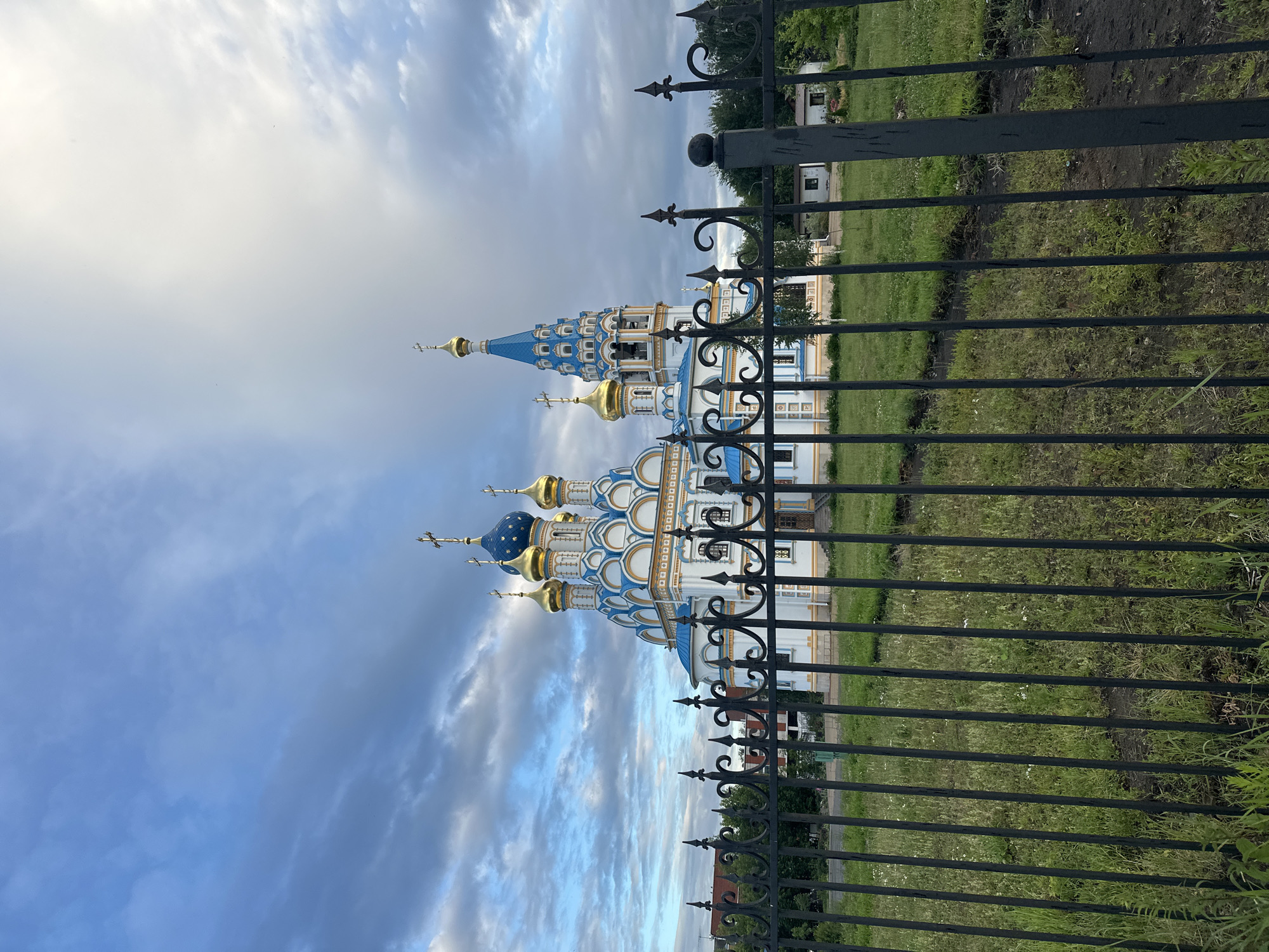 The image shows a blue and white domed church with gold crosses, framed by a black metal fence. The church has intricate architectural details, including ornate window frames and a tall, tapering blue spire. The sky is partly cloudy with patches of blue and grey clouds, suggesting it might be late afternoon or early evening. The fence appears to be made of wrought iron with decorative spikes and curves. Behind the church, there is a green grassy area with some trees and a small building, possibly a house or another part of the church complex. The overall scene conveys a sense of religious and architectural grandeur against a natural backdrop. There are no people visible in the image, and no text is discernible. Given the location context of Moscow, Russia, this is likely a Russian Orthodox church.